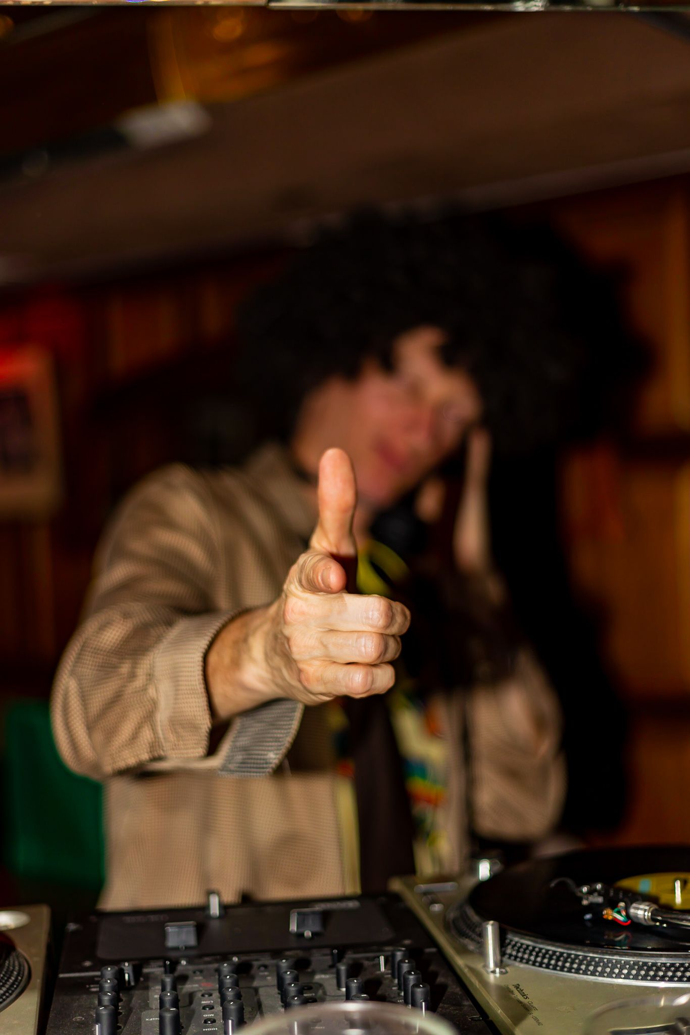 A DJ in an afro wig points toward the camera while standing behind turntables in a warmly lit venue.