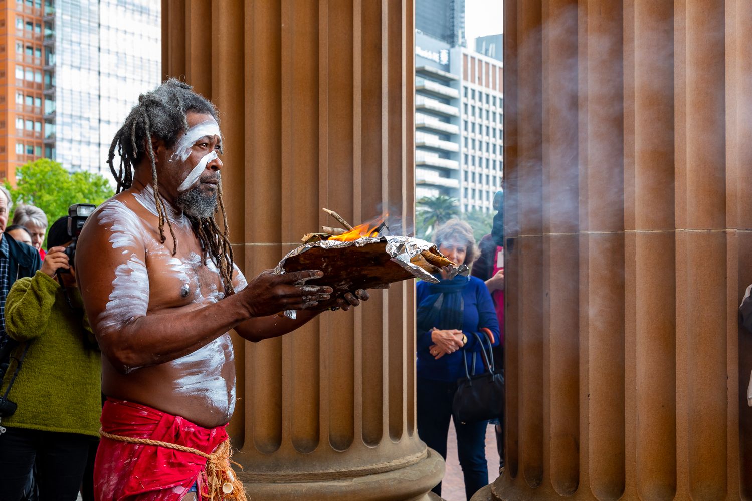 Aboriginal man with body paint holds a smoking ceremony bowl beside sandstone columns while onlookers watch in a city setting.