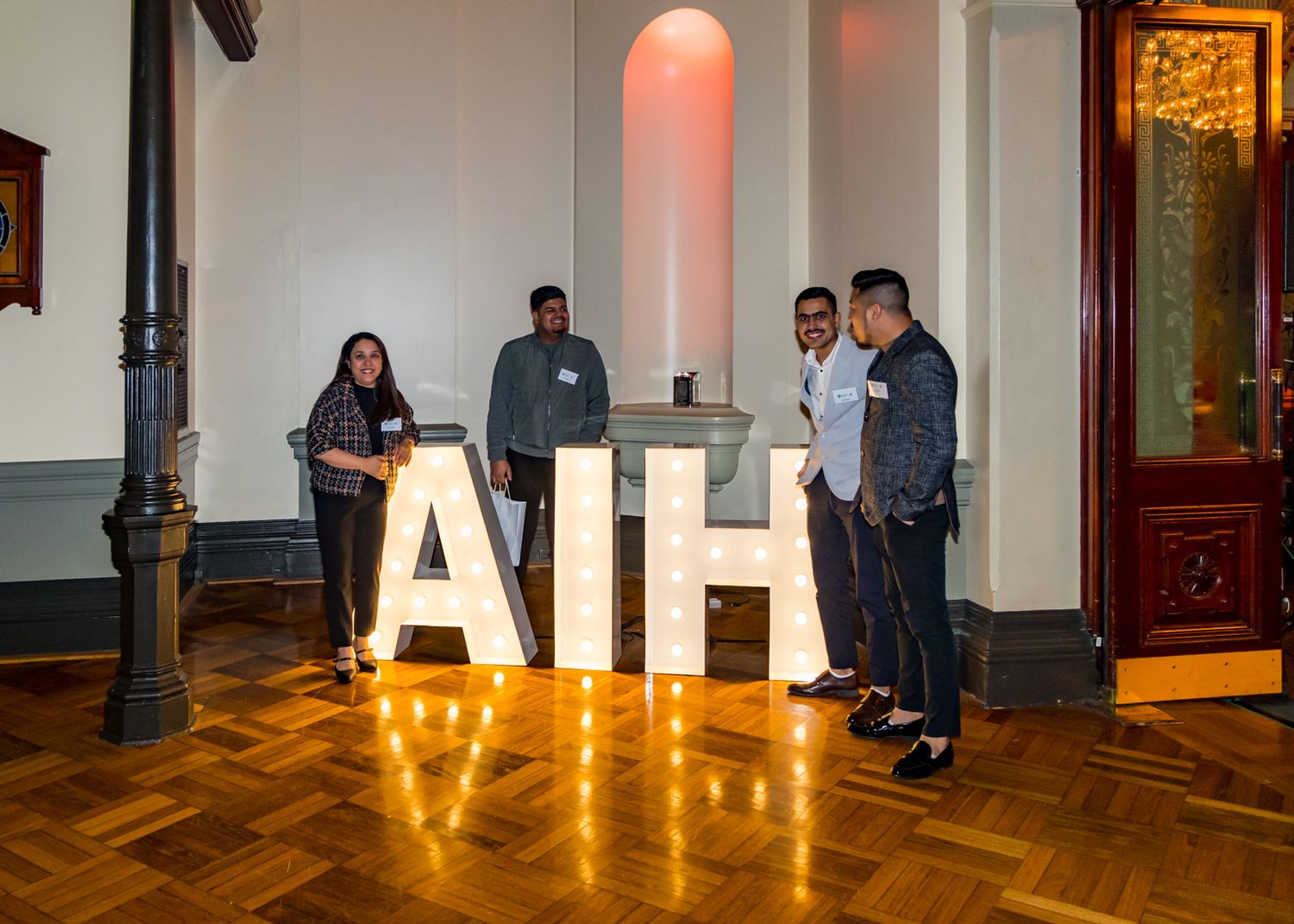 Four people stand beside large illuminated marquee letters spelling AIH on a wooden floor in an elegant historic hall.