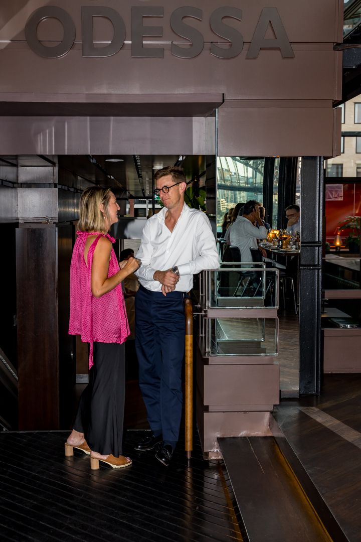 A man in a white shirt and a woman in a pink top chat at the entrance of the Odessa restaurant, with diners seated inside.