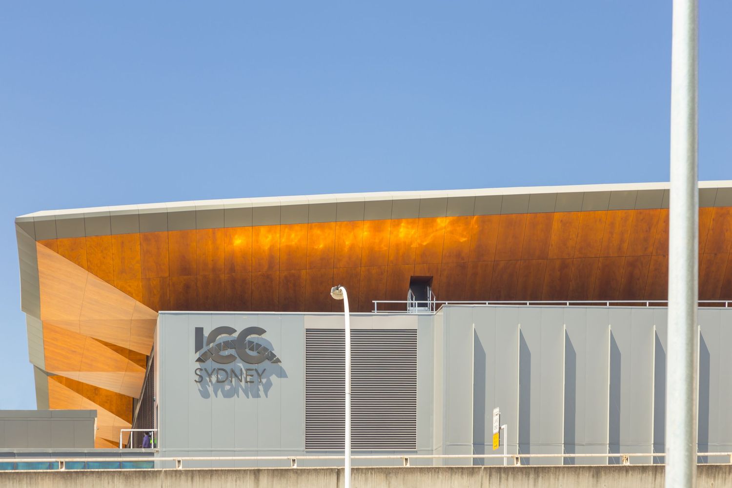 ICC Sydney building exterior with glowing copper-toned timber roof curve, grey cladding and logo signage against a clear blue sky.