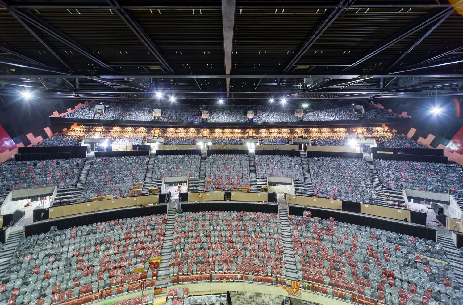 Wide view of an indoor arena showing circular tiered seating, upper level suites, and steel roof structure with bright overhead lighting.