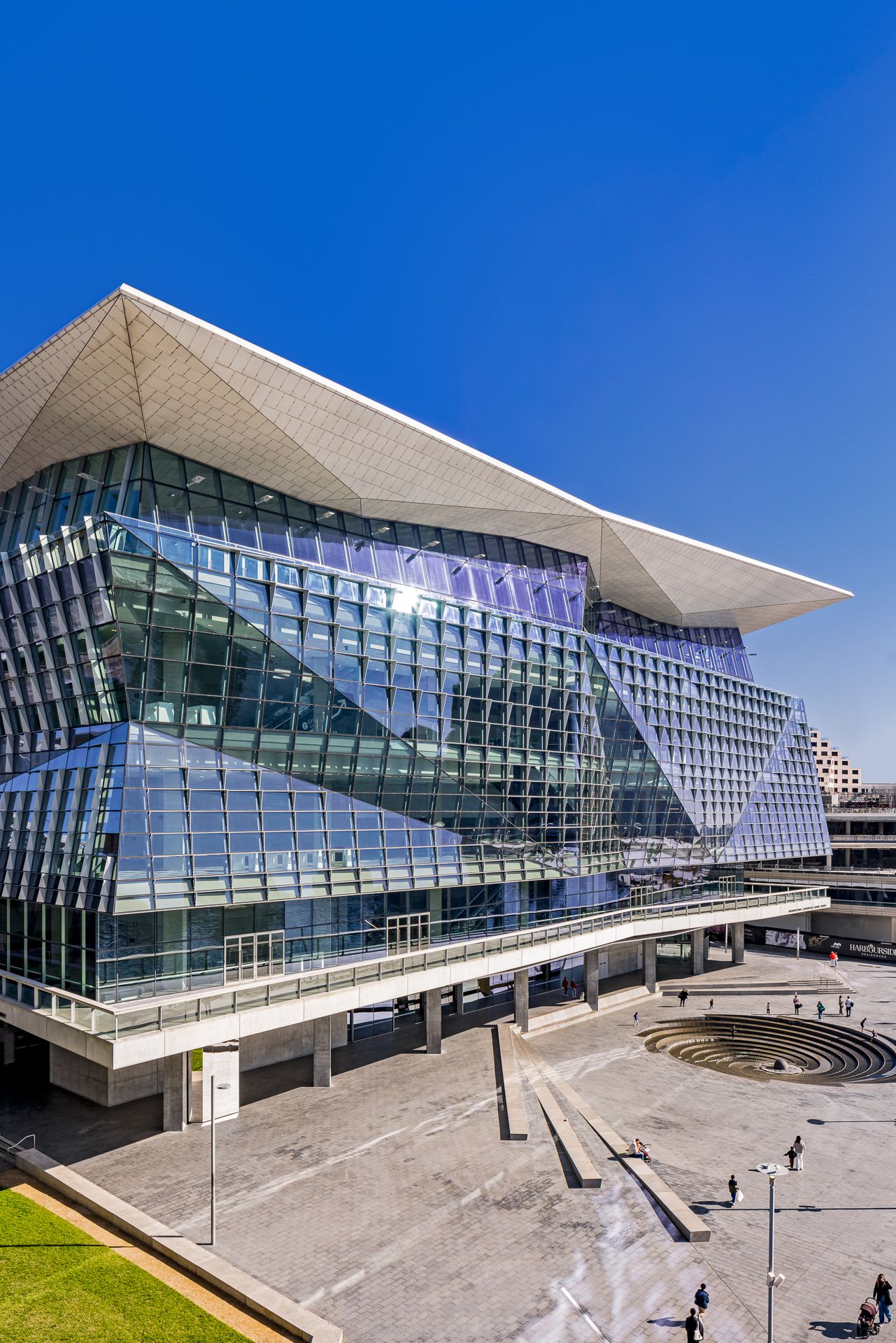 Geometric glass convention building with a dramatic angular roof and a circular sunken plaza where visitors walk in sunshine.  ICC Sydney