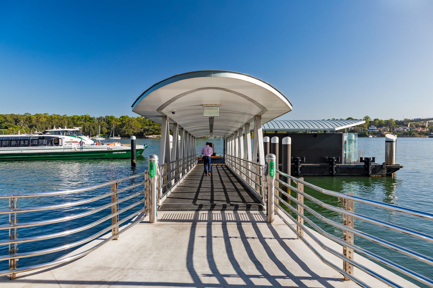 Person walking along a covered gangway toward a ferry wharf with metal railings, with a ferry moored beside it on sunny water.
