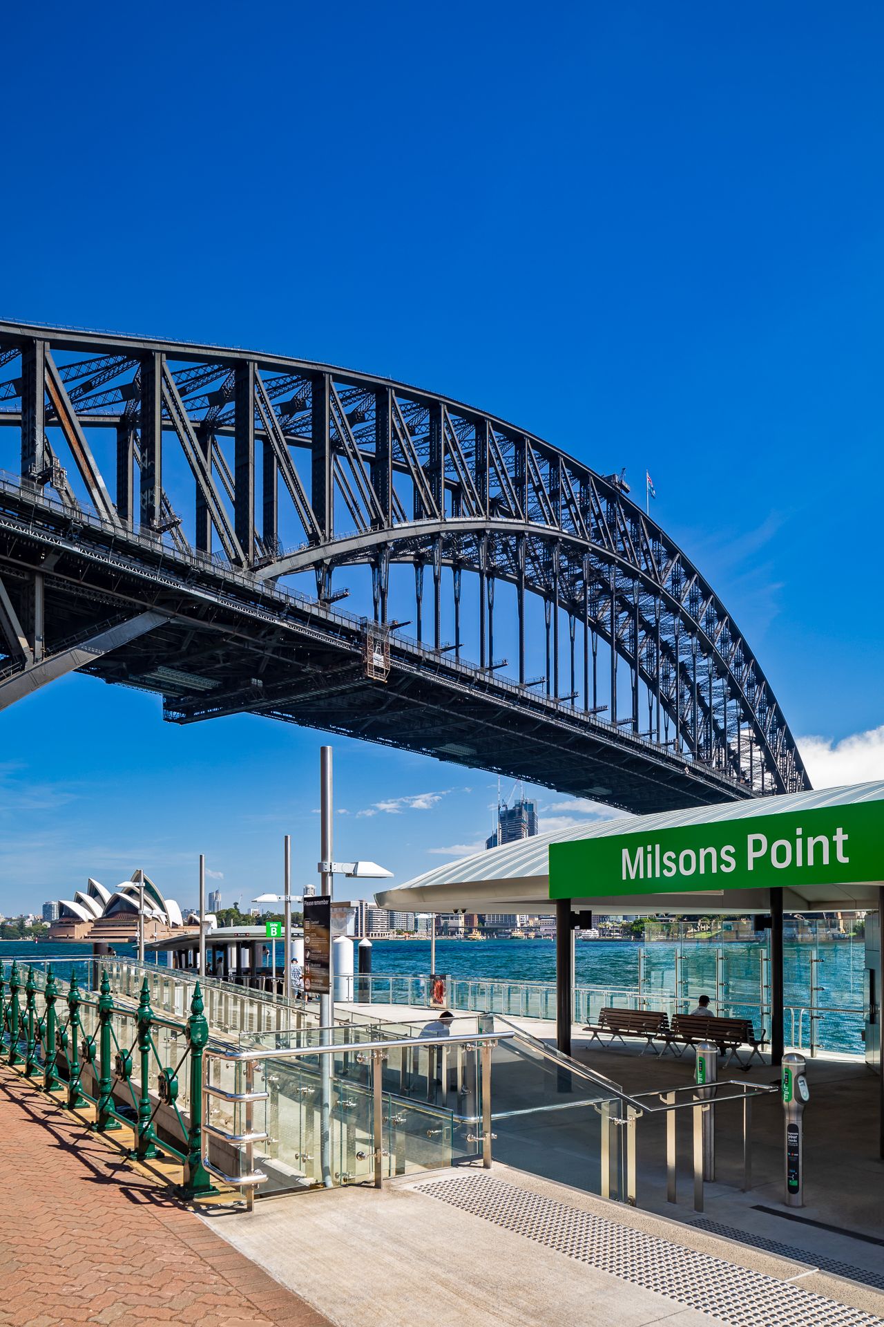 Milsons Point ferry wharf sign beneath the Sydney Harbour Bridge, with the Opera House visible across the sunny blue harbour.