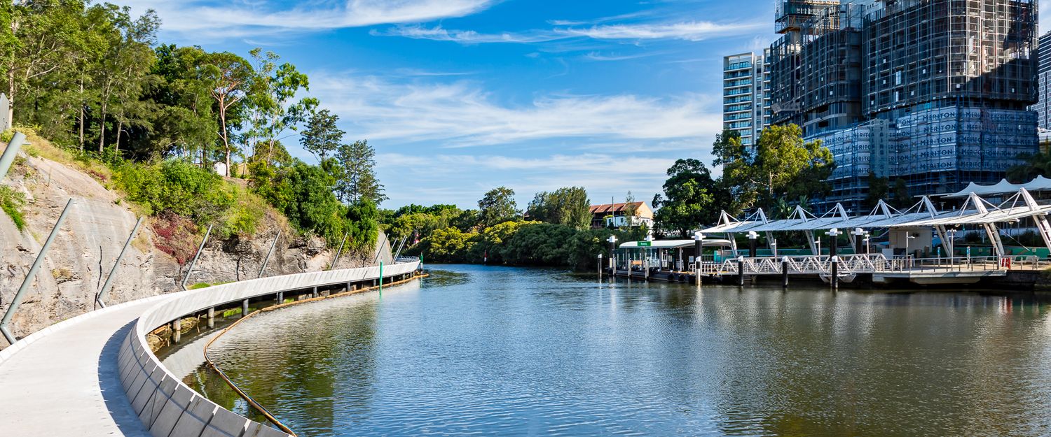 Curved waterfront walkway beside a calm river, lined with trees and a rock face, with apartment buildings and a wharf ahead.