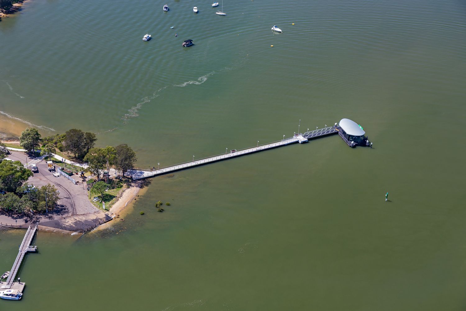Aerial view of a long narrow pier extending from a sandy beach to a round-roofed ferry wharf, with boats scattered across calm green water.