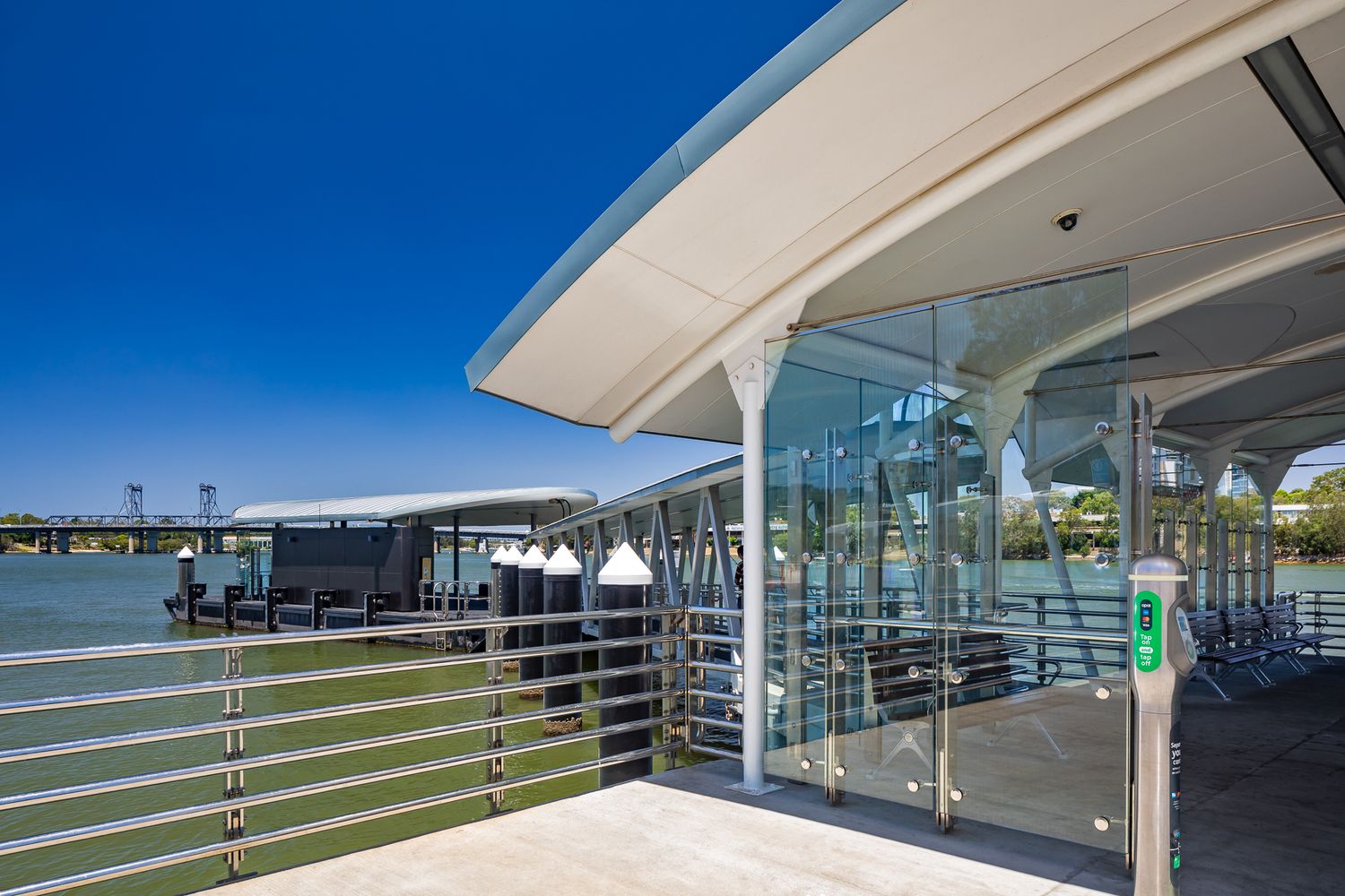 Close-up of a contemporary ferry wharf with a sweeping white canopy, glass panels and metal railings overlooking calm green water.