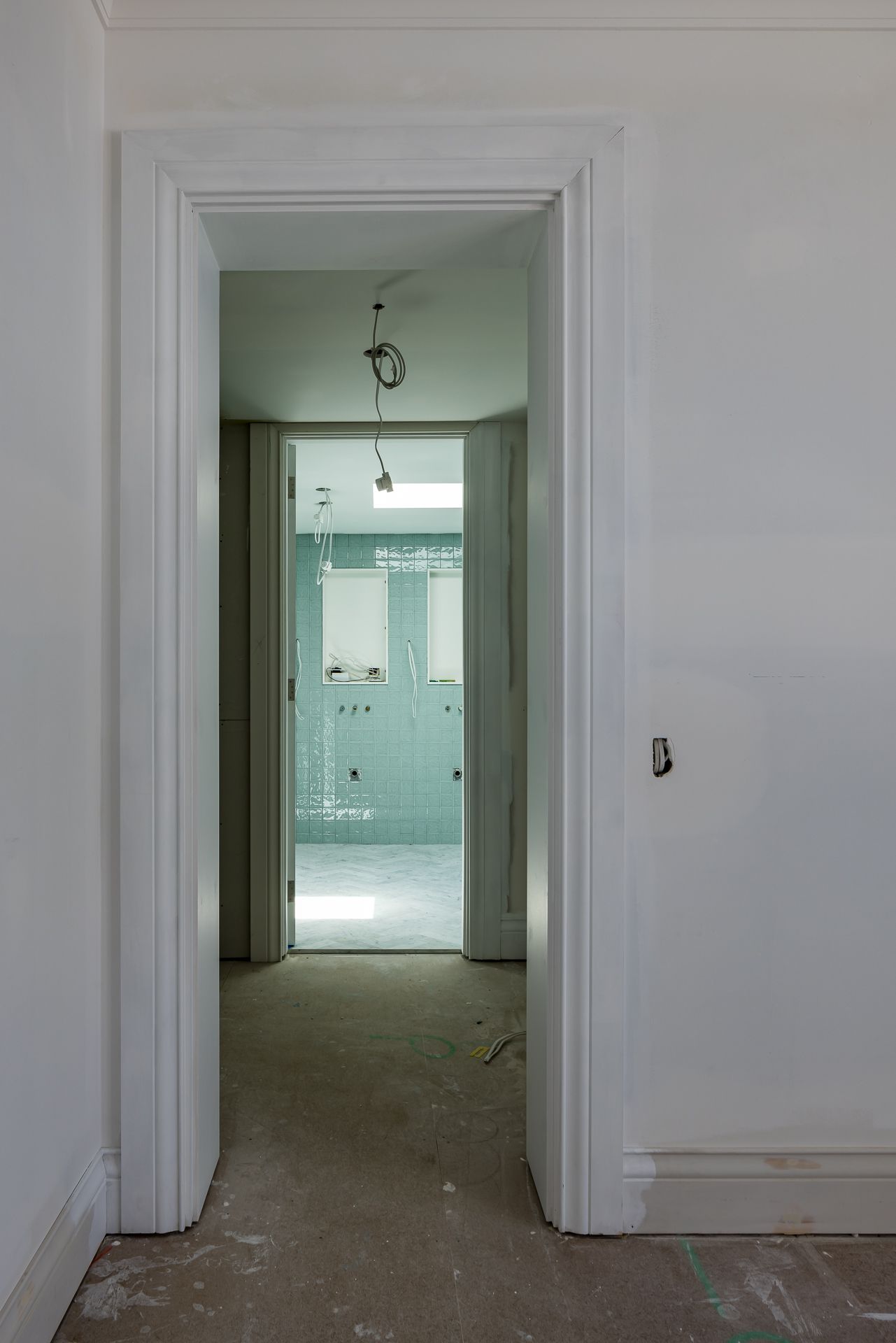 View through a white doorway into a renovated bathroom with soft green tiles and a skylight, beyond a bare unfinished floor.