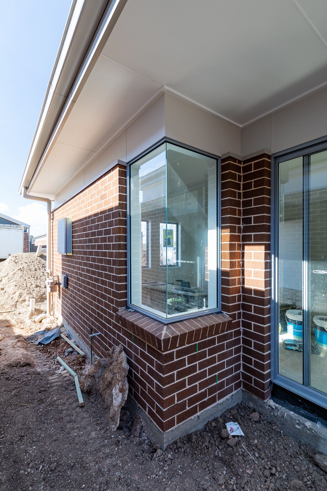 Corner of a new red brick home with large windows, a sliding door and eaves, surrounded by bare soil during construction.