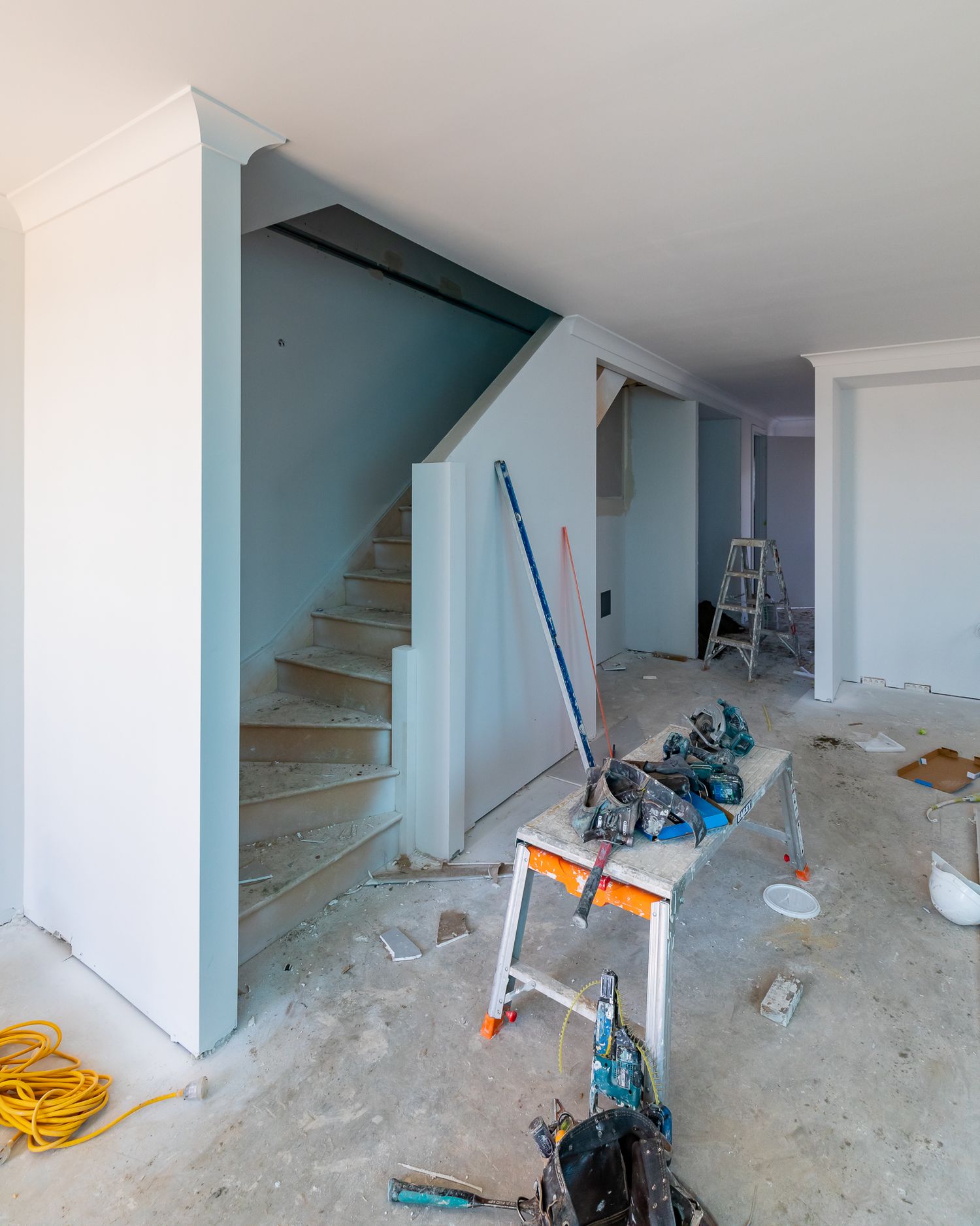 Unfinished home interior with a staircase, bare plaster walls, a work bench, ladder and scattered tools on the floor.