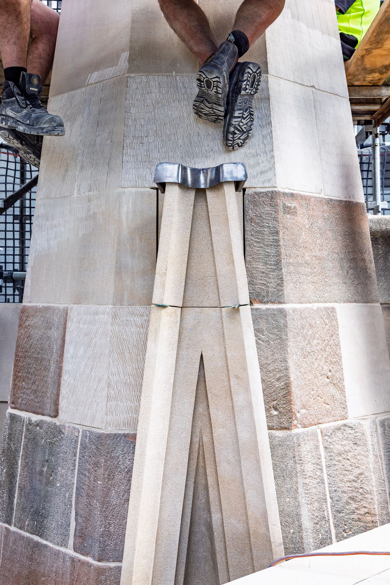 Worker's legs in hiking boots hang over a freshly carved sandstone pinnacle base with a metal clamp during restoration.