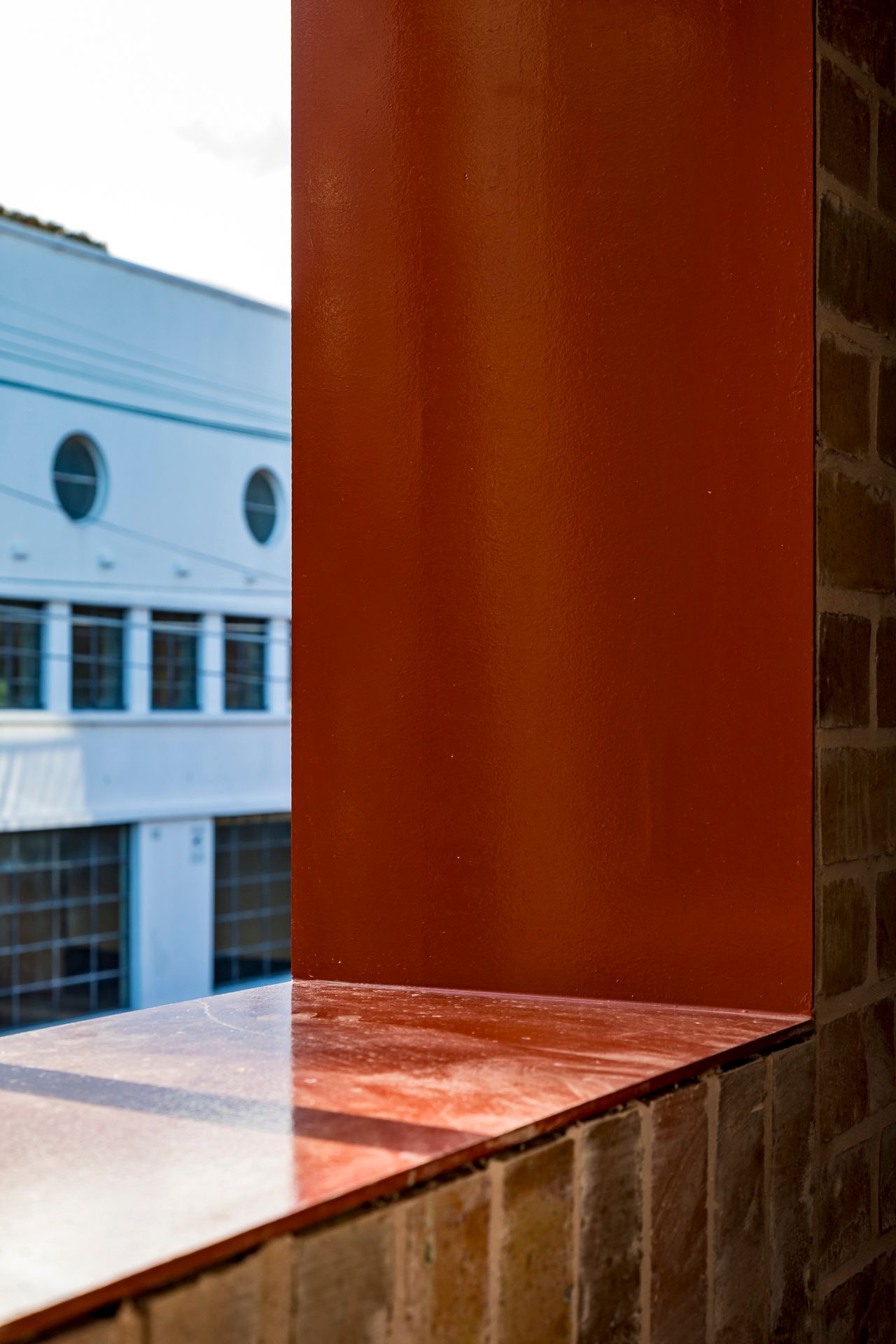 Red steel window frame and glossy sill set into a brick wall with a white building visible outside.