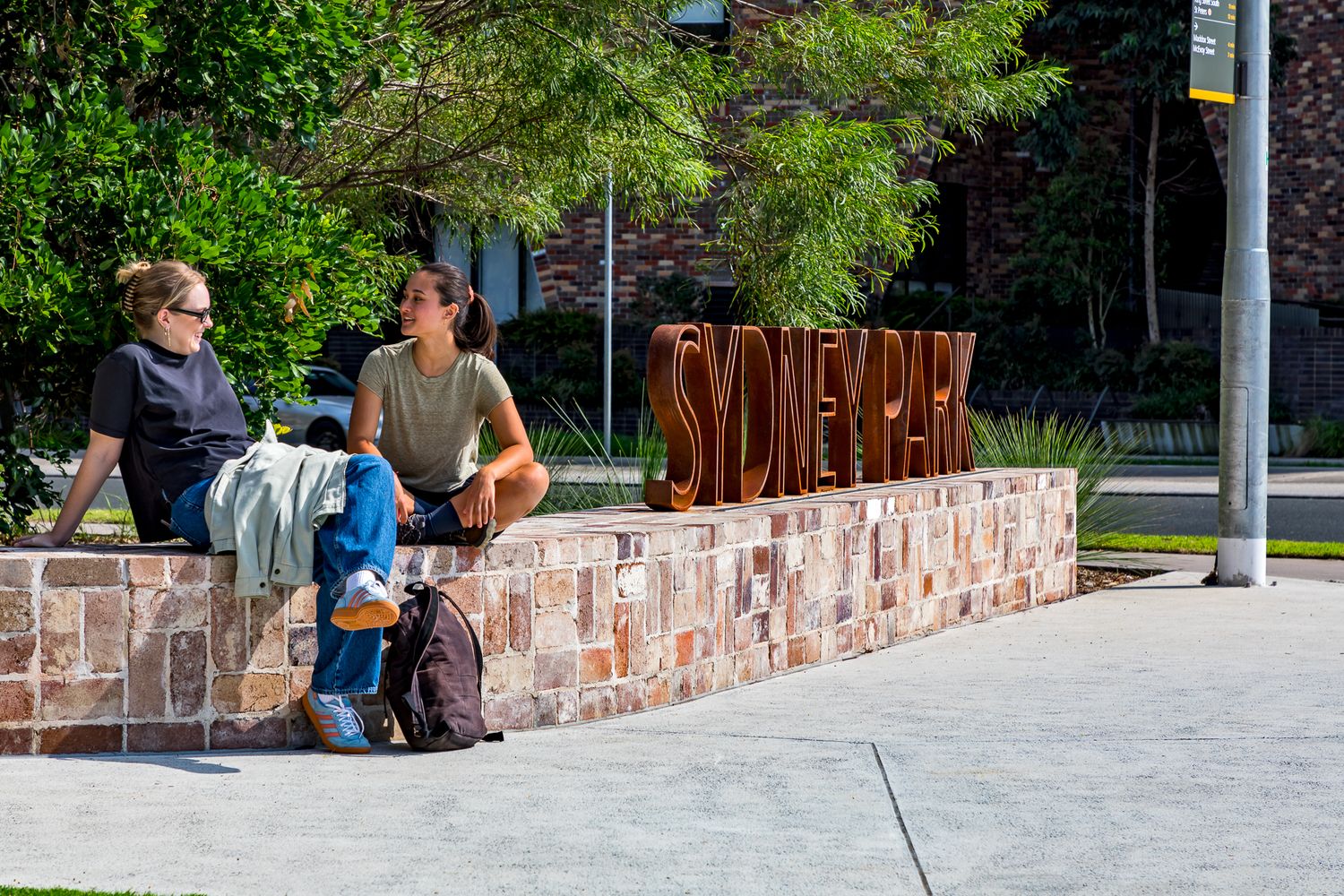 Two friends sit chatting on a sandstone block wall next to large rusty steel Sydney Park letters.