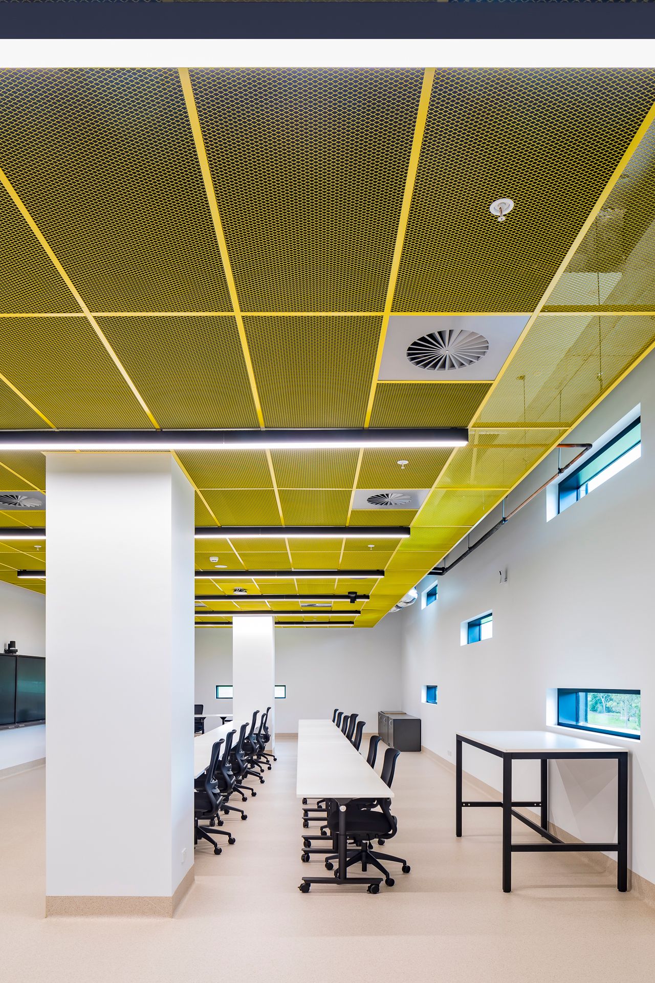 Bright classroom with yellow mesh ceiling panels, white columns, long tables and black rolling chairs.