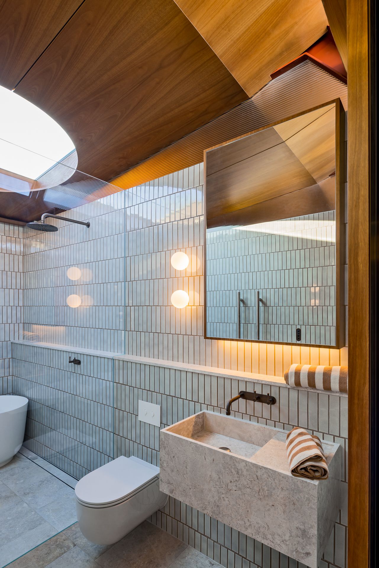 Modern bathroom with vertical gray tiles, timber ceiling, skylight, stone sink, wall-mounted toilet, freestanding tub, and brass fixtures.