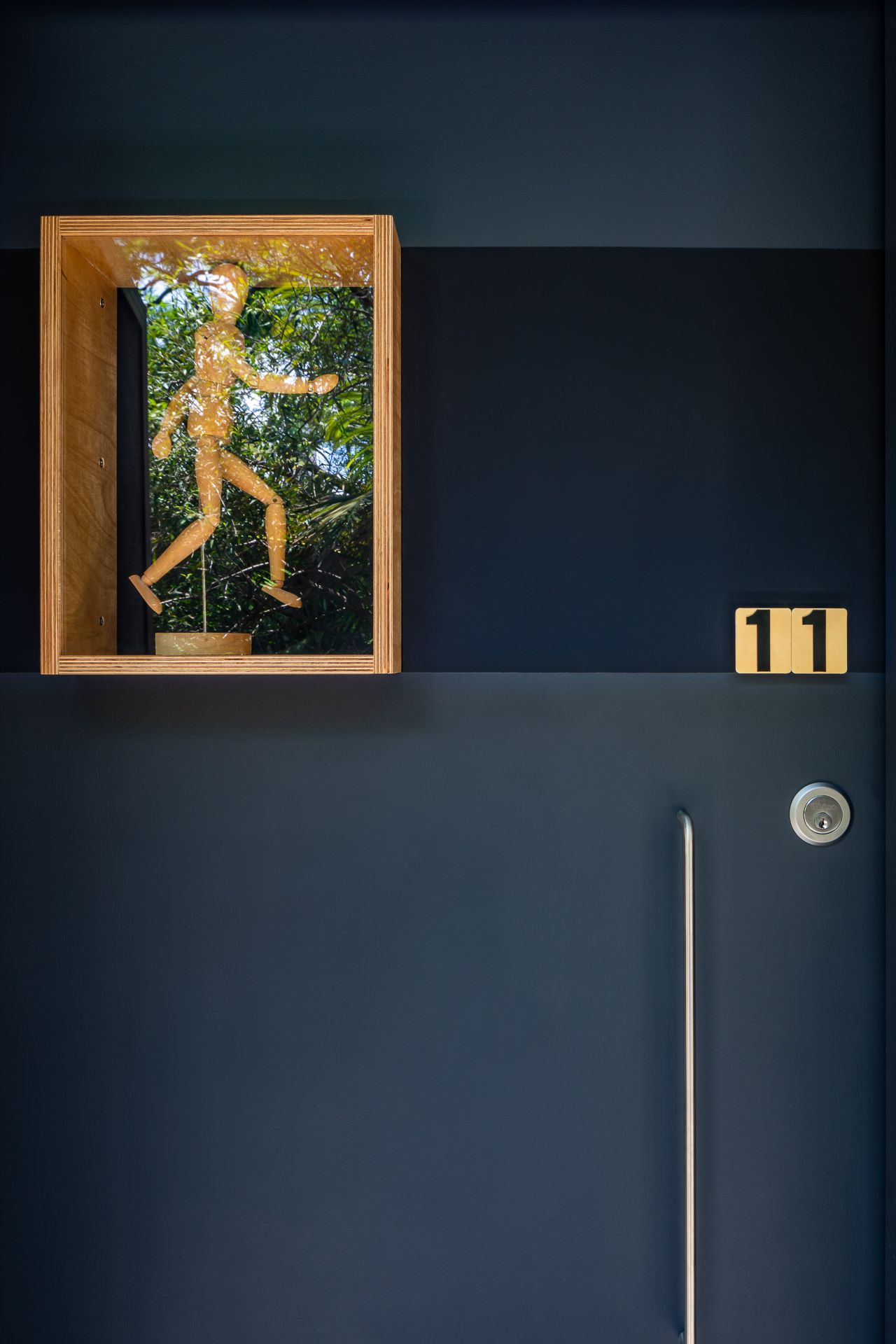 Dark navy apartment door numbered 11 with a wooden display box holding a small wooden figure against green foliage.
