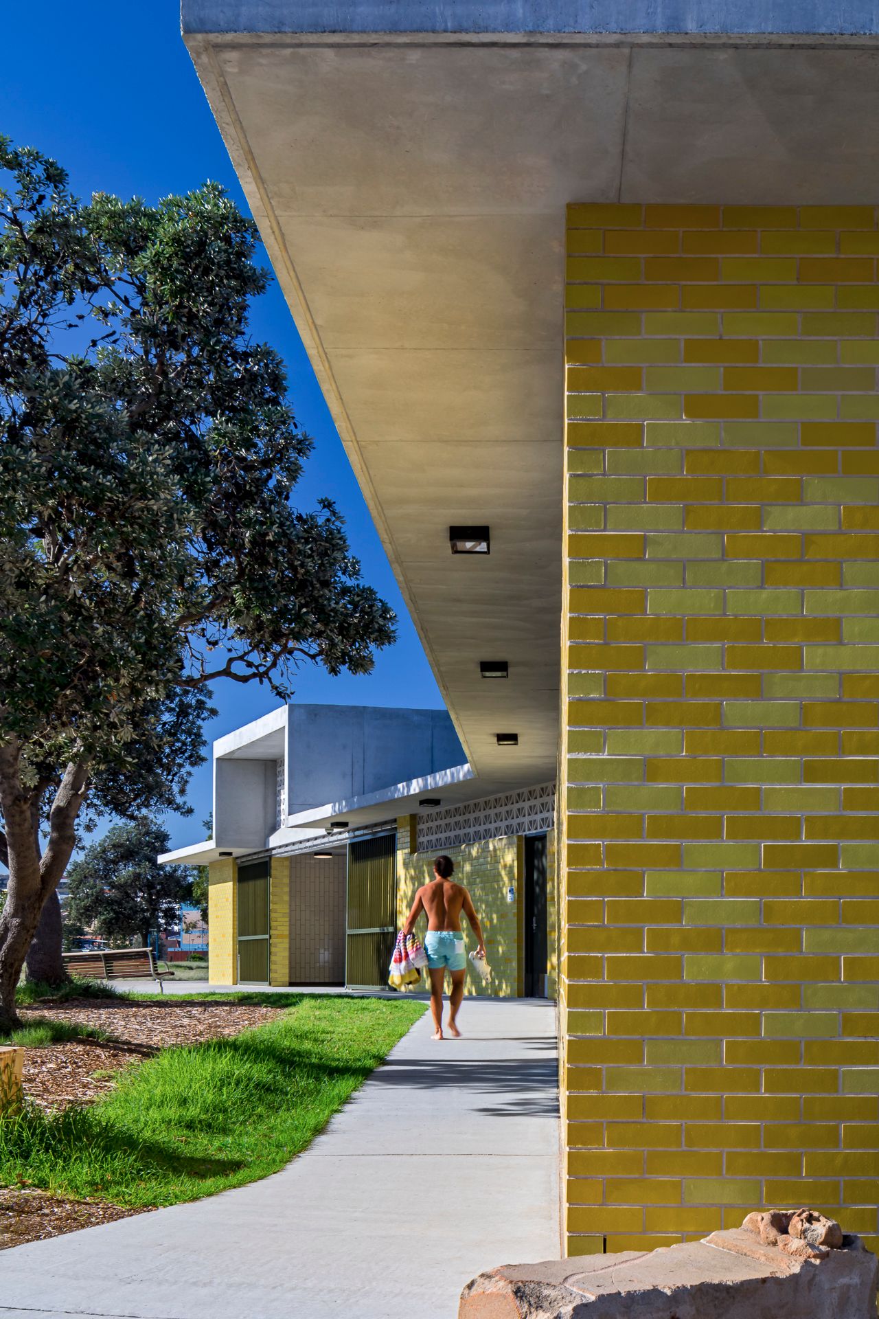 A person in swim shorts walks along a concrete path beside a yellow brick building with a cantilevered concrete roof and a shady tree.