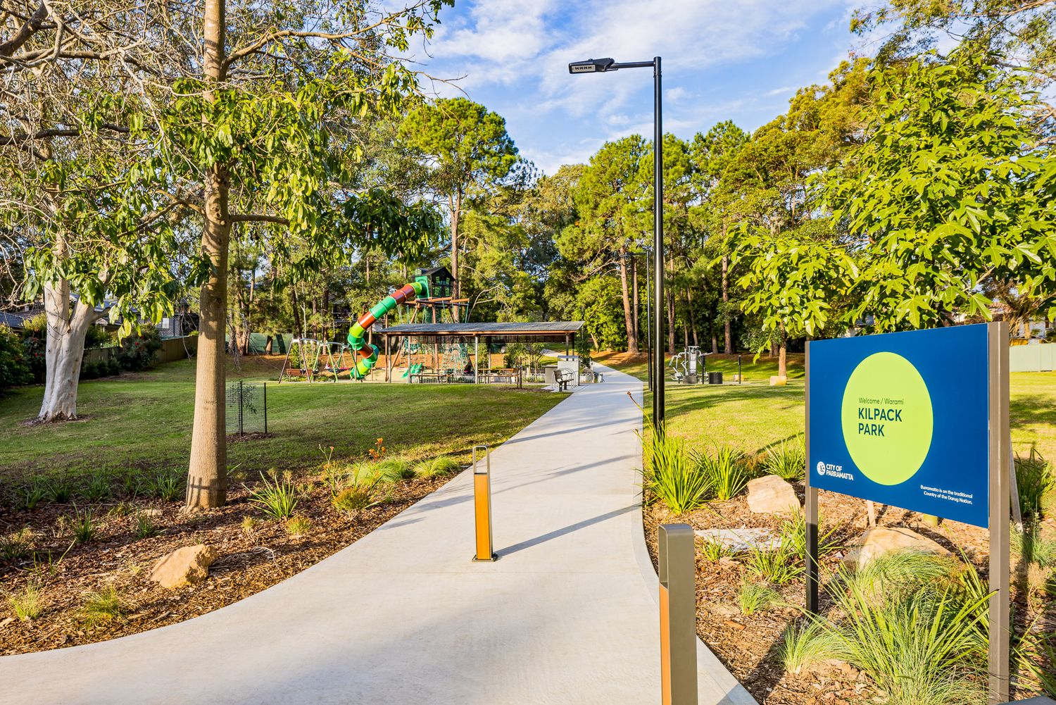 Concrete park pathway leads toward a playground and shelter, with a blue park entrance sign, lamp post and native garden beds.