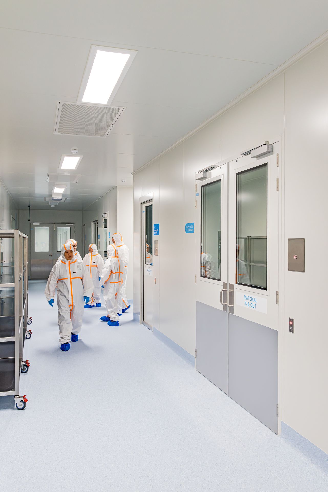 Technicians in protective suits and blue shoe covers walk down a bright cleanroom corridor lined with double doors.