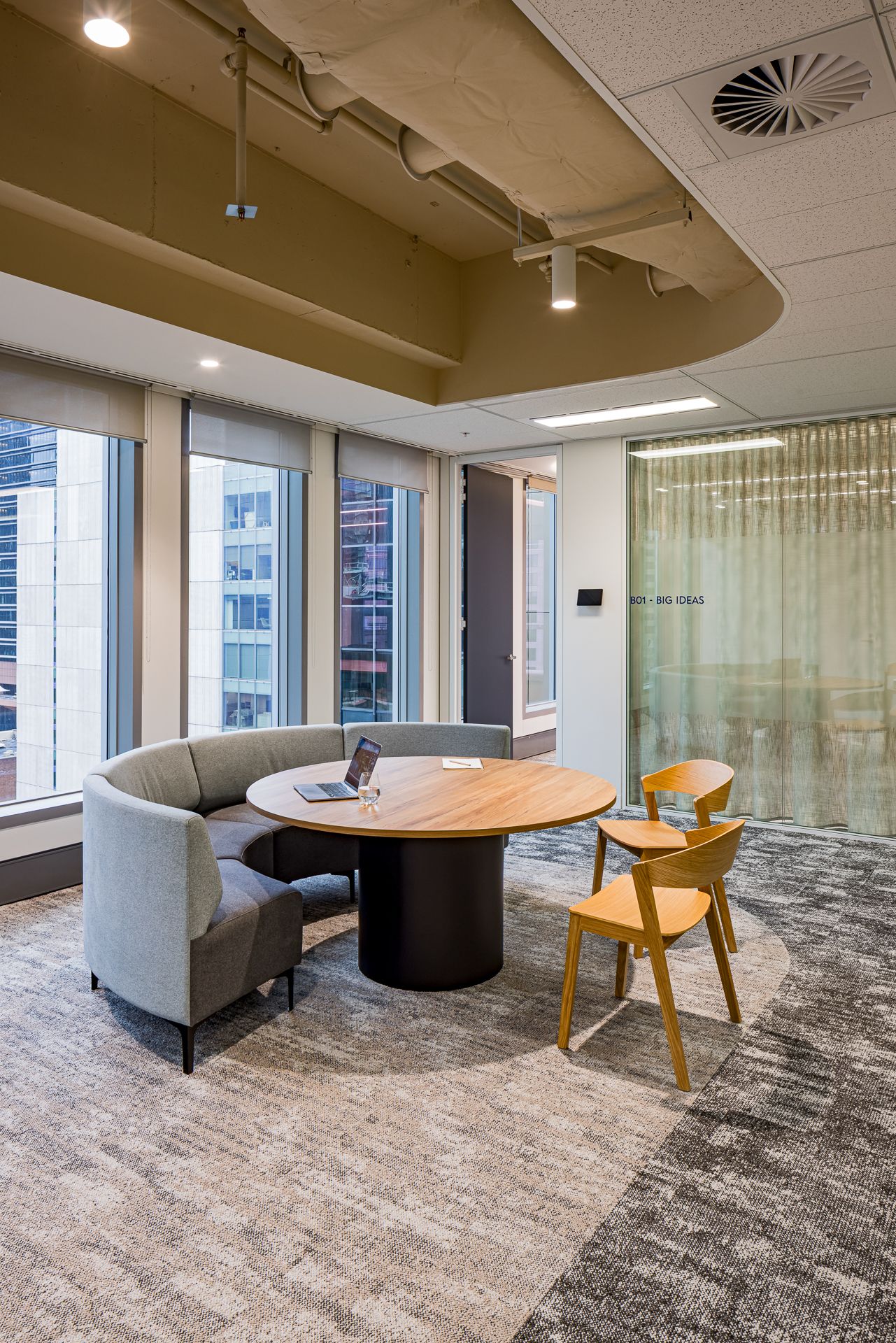Office breakout area with a curved grey banquette, round timber table and orange chairs beside windows overlooking the city.