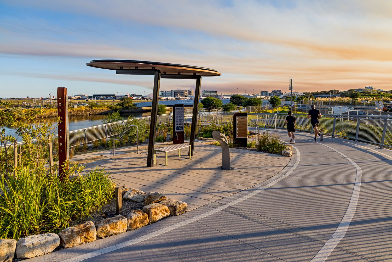 Joggers on a curved waterfront path beside a circular shade structure, benches, interpretive signs and native grasses at sunset.
