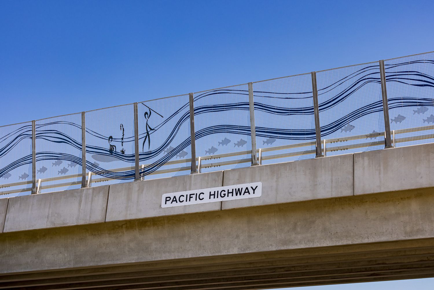 Bridge barrier with wave-patterned artwork above a Pacific Highway sign against a bright blue sky.