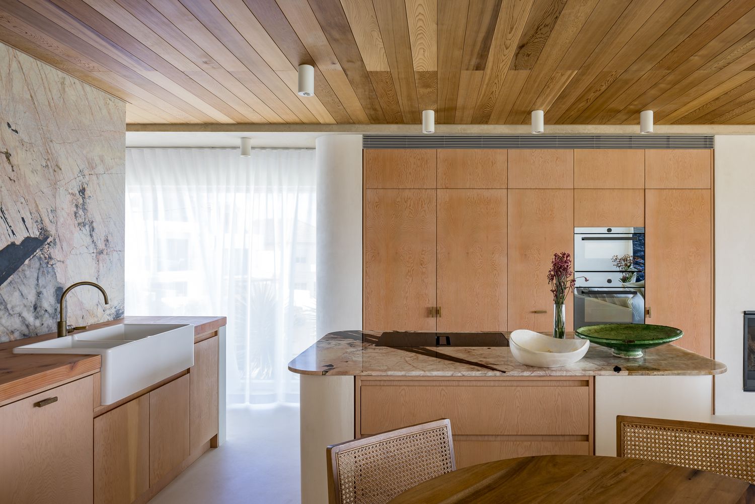 Modern kitchen with timber cabinetry, a stone-topped island, farmhouse sink and a warm wood plank ceiling beside sheer curtains.