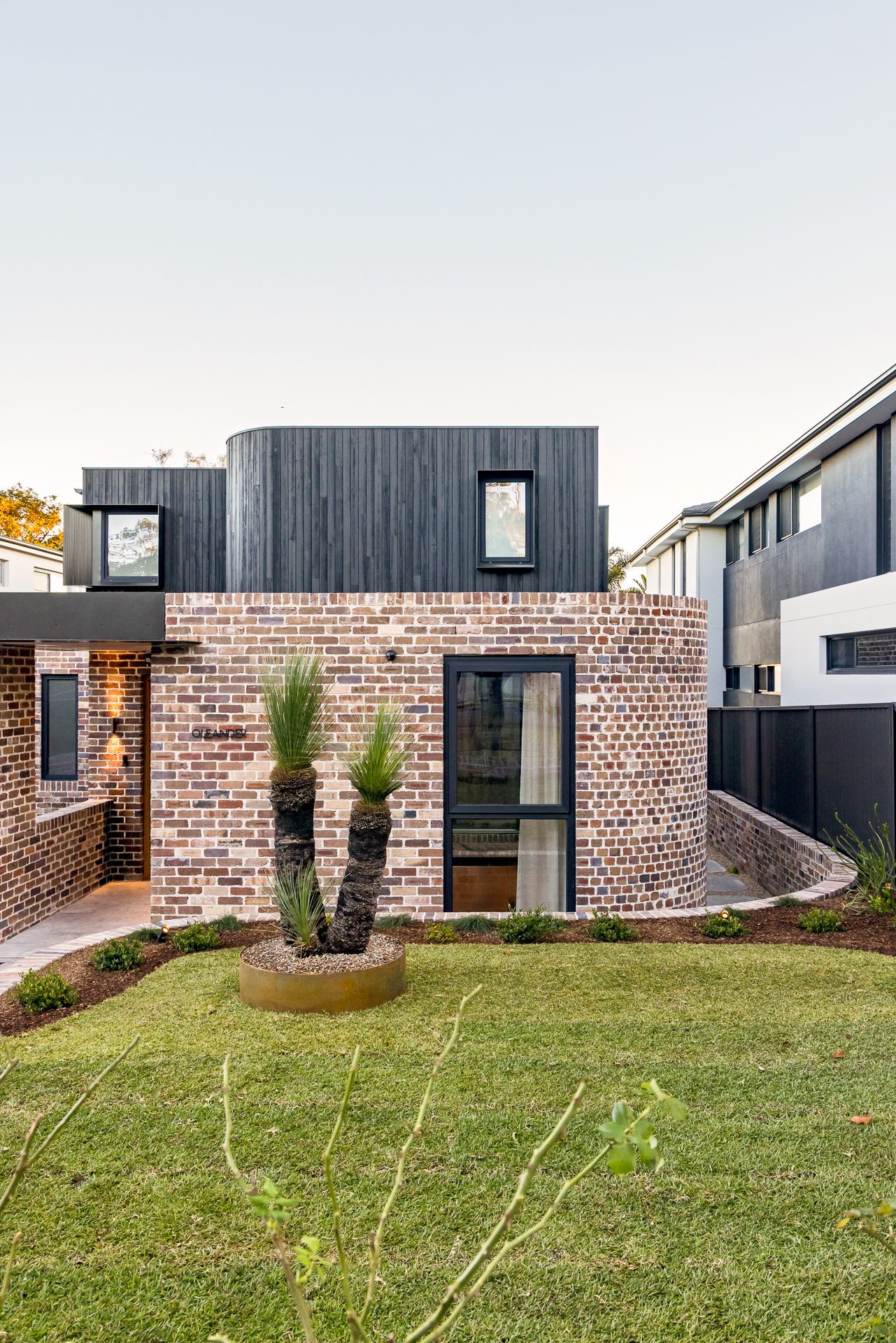 Modern brick house with a dark timber upper level, a curved brick wall, grass trees in a round planter and a green lawn.