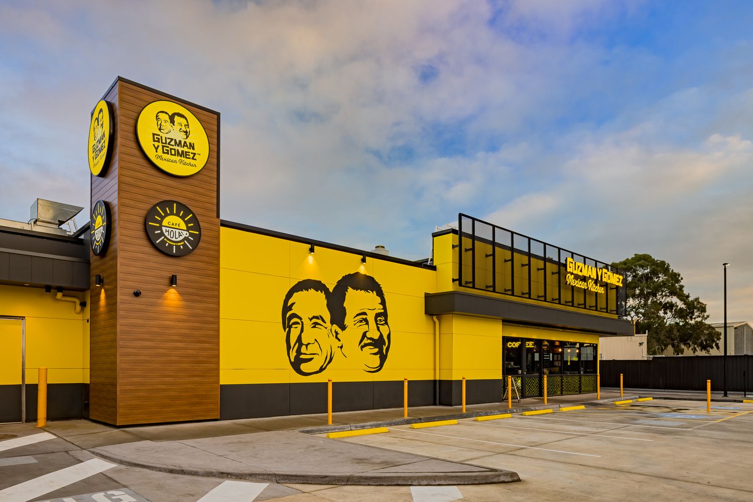 Corner view of a yellow Guzman y Gomez restaurant featuring a tall brick sign tower and a large mural of two smiling men.