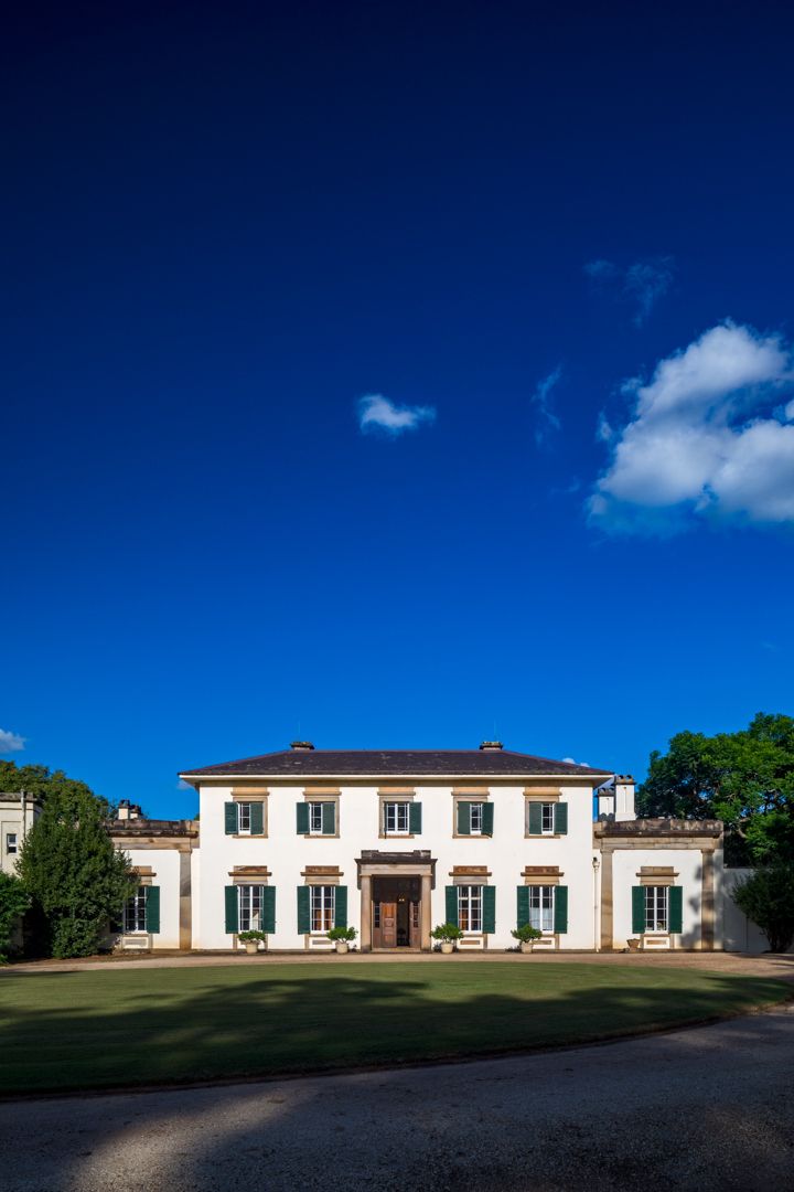 Symmetrical white two-storey colonial mansion with green shutters and a central entrance, set behind a manicured lawn under a deep blue sky.