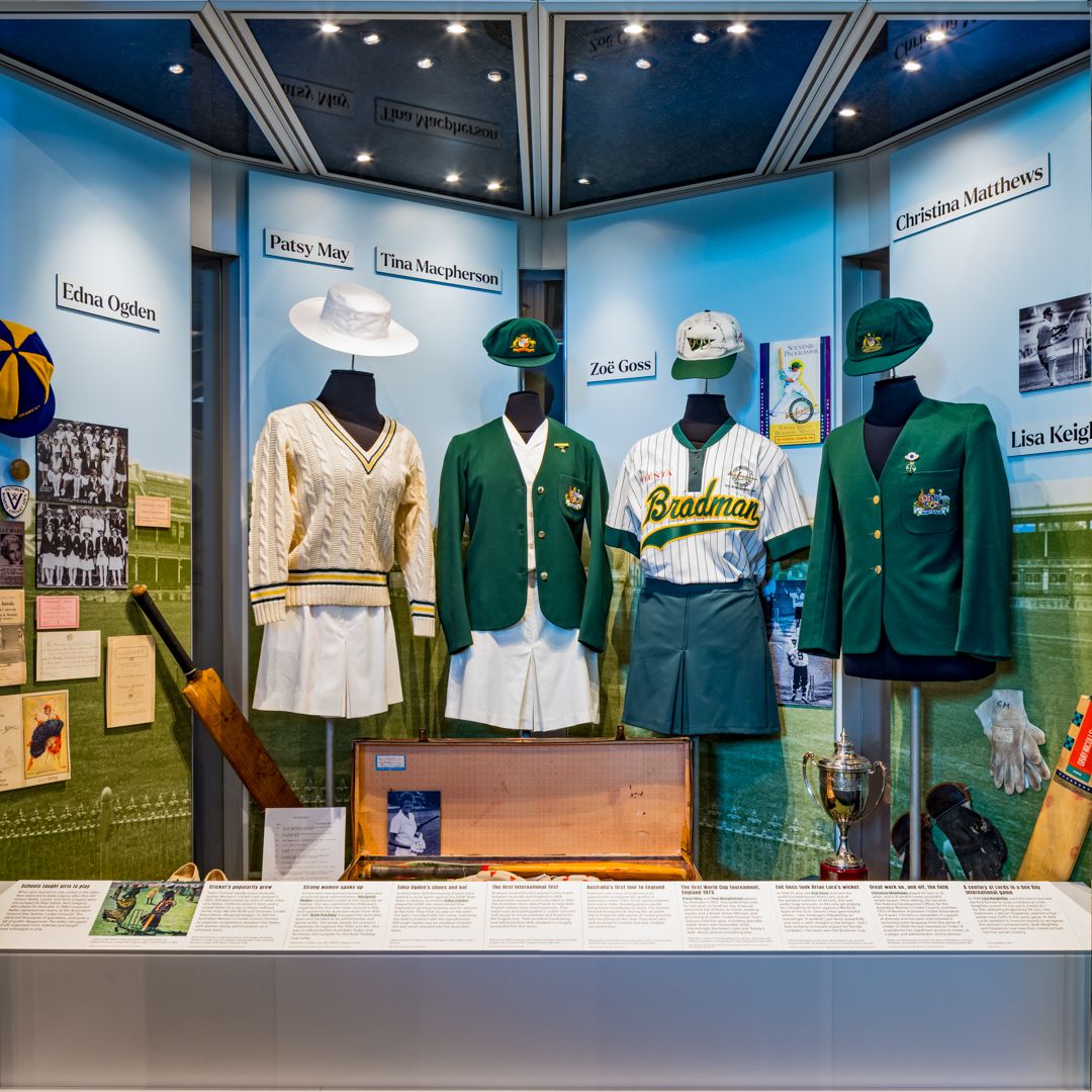 Museum display of women's cricket uniforms on mannequins, including a green Baggy Green blazer and a Bandman jersey, with trophies.