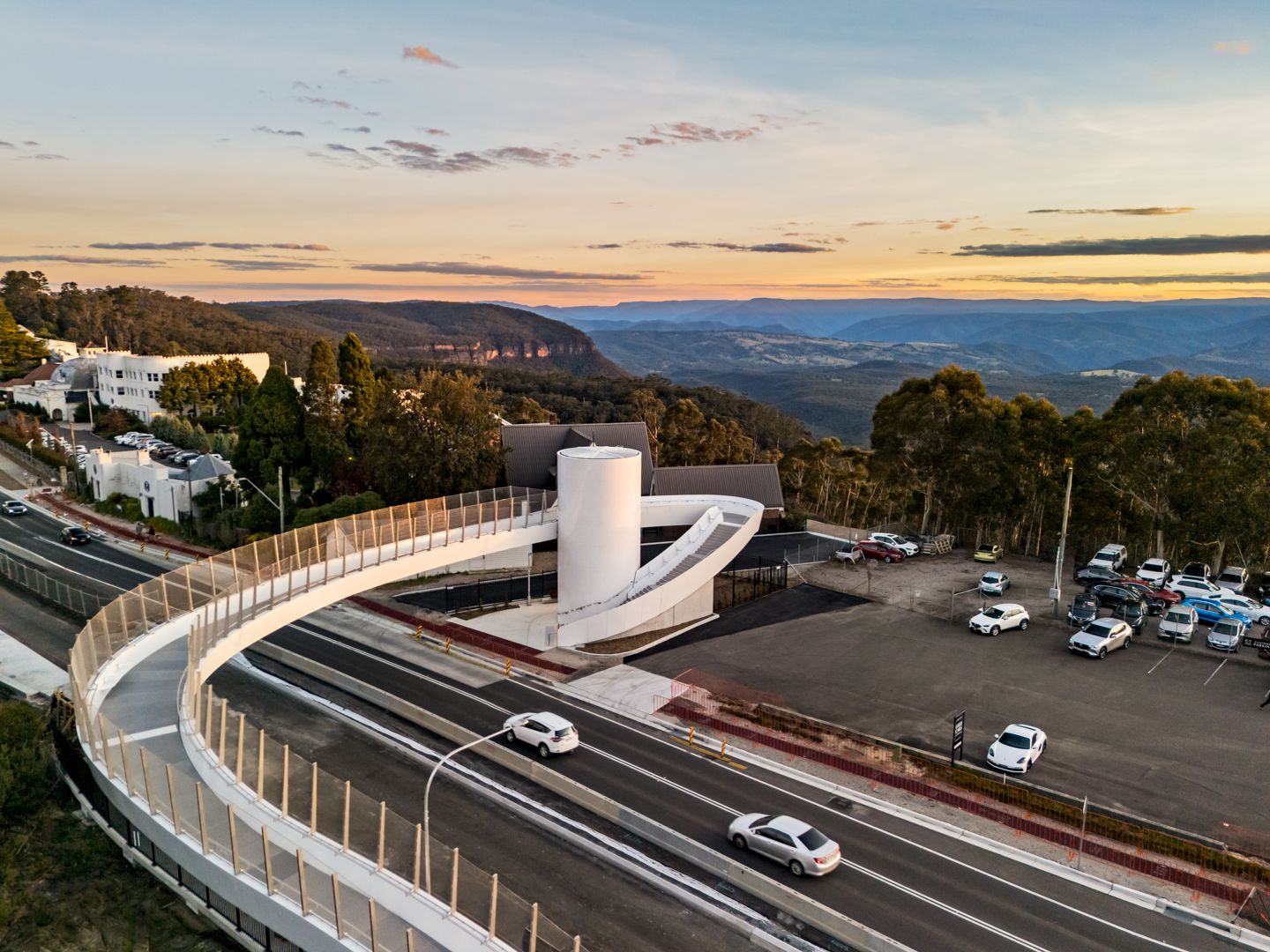 Modern curved pedestrian footbridge and white cylindrical tower beside a road and car park, overlooking forested valleys at sunset.