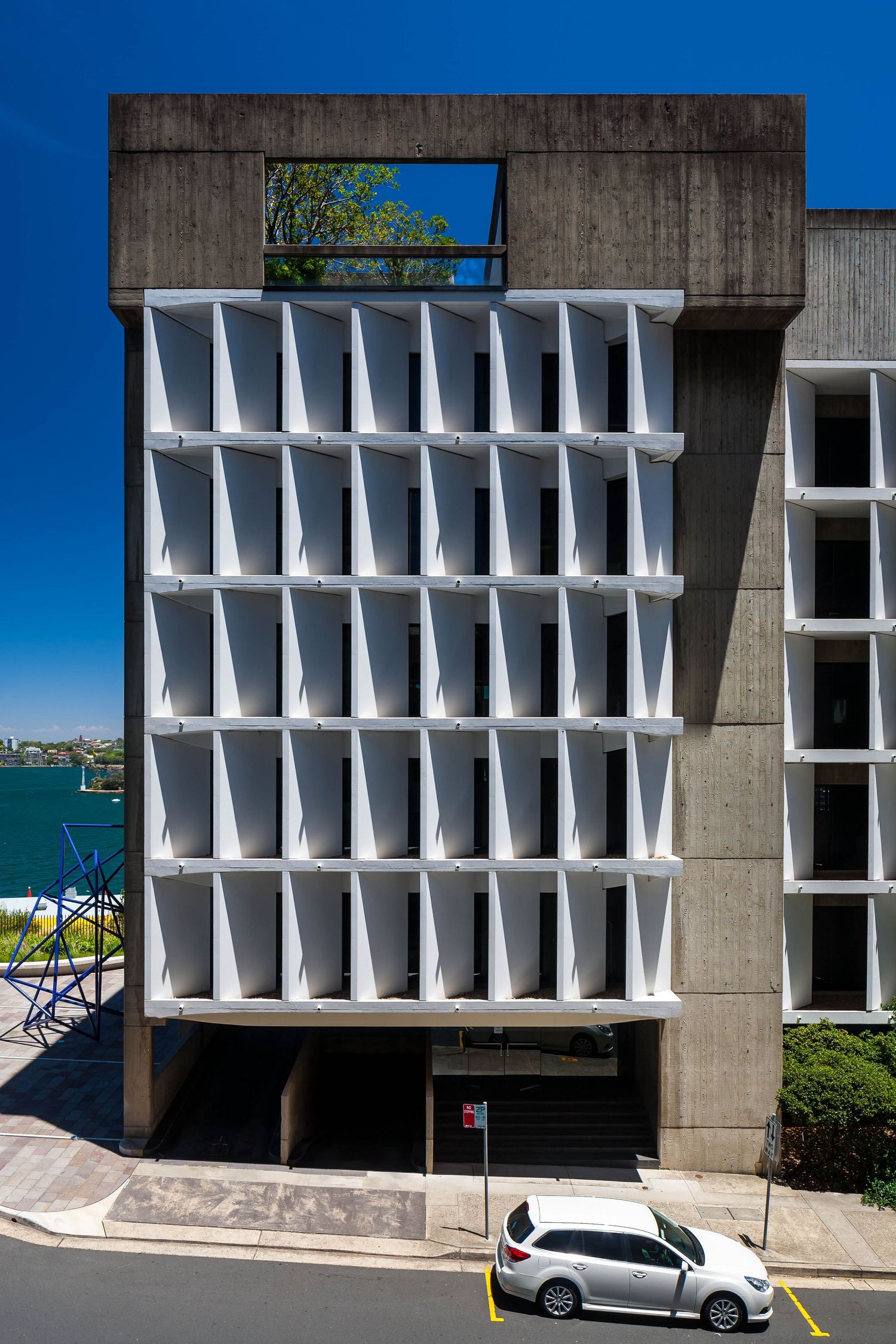 Brutalist concrete office building with white louvred facade panels overlooking blue harbour waters on a clear sunny day.