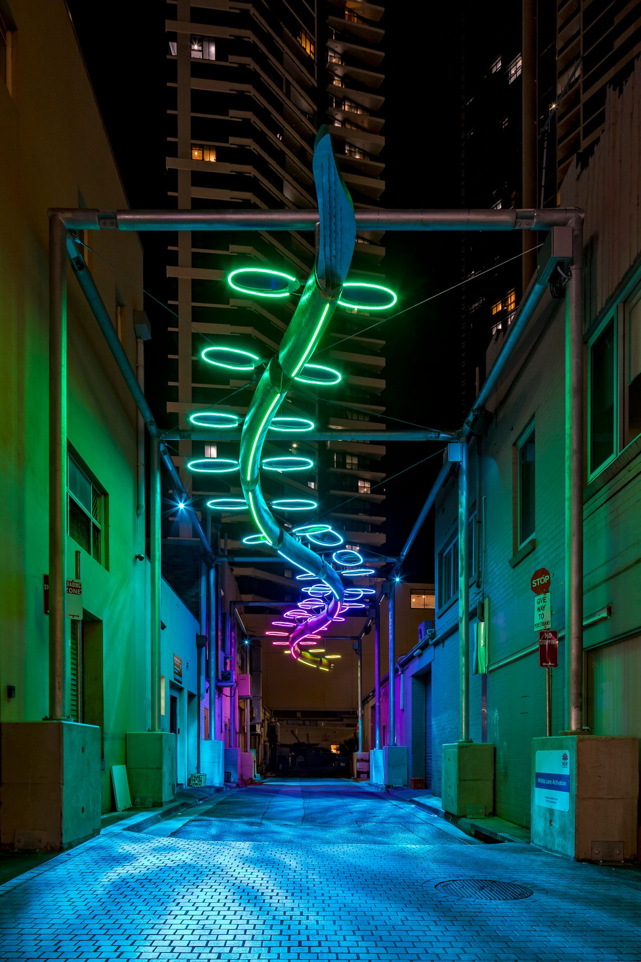 Green neon rings spiral above a cobblestone city alley, lighting the walls and surrounding high-rises at night.