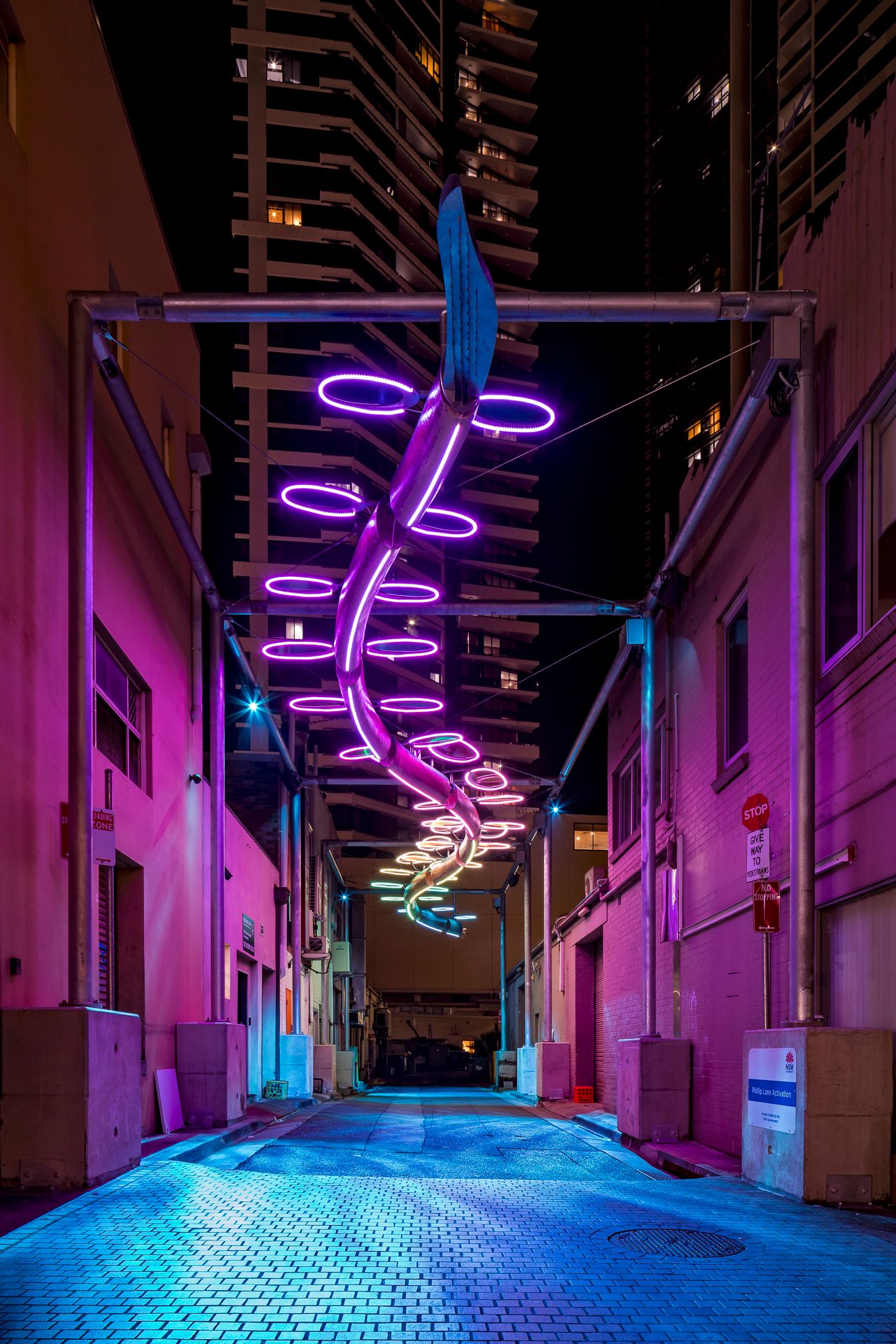 Violet neon rings twist along a suspended sculpture above a quiet downtown alley lit in magenta and blue.