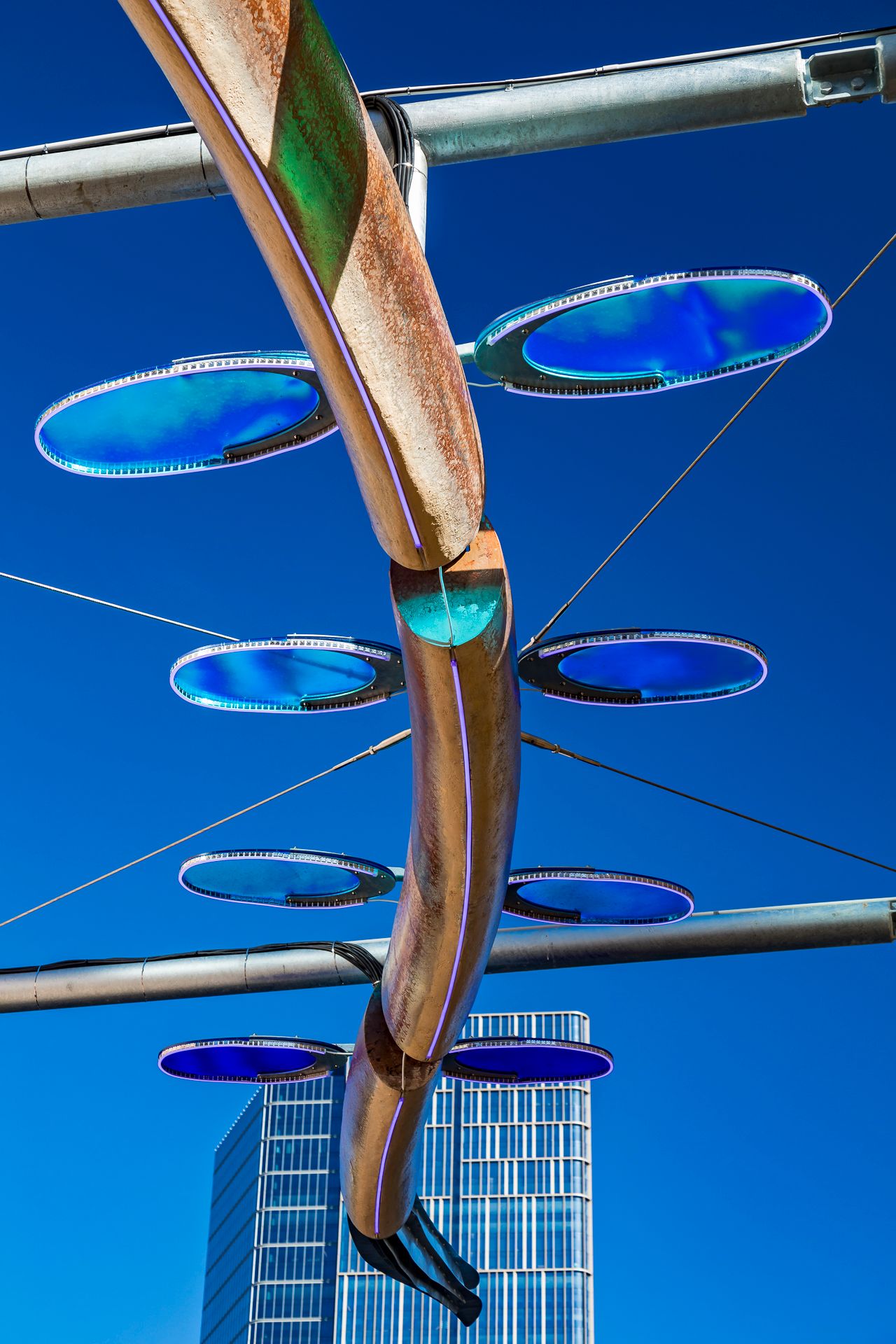 Bronze curved sculpture with blue translucent oval discs suspended from steel beams against a vivid blue sky and a glass skyscraper.