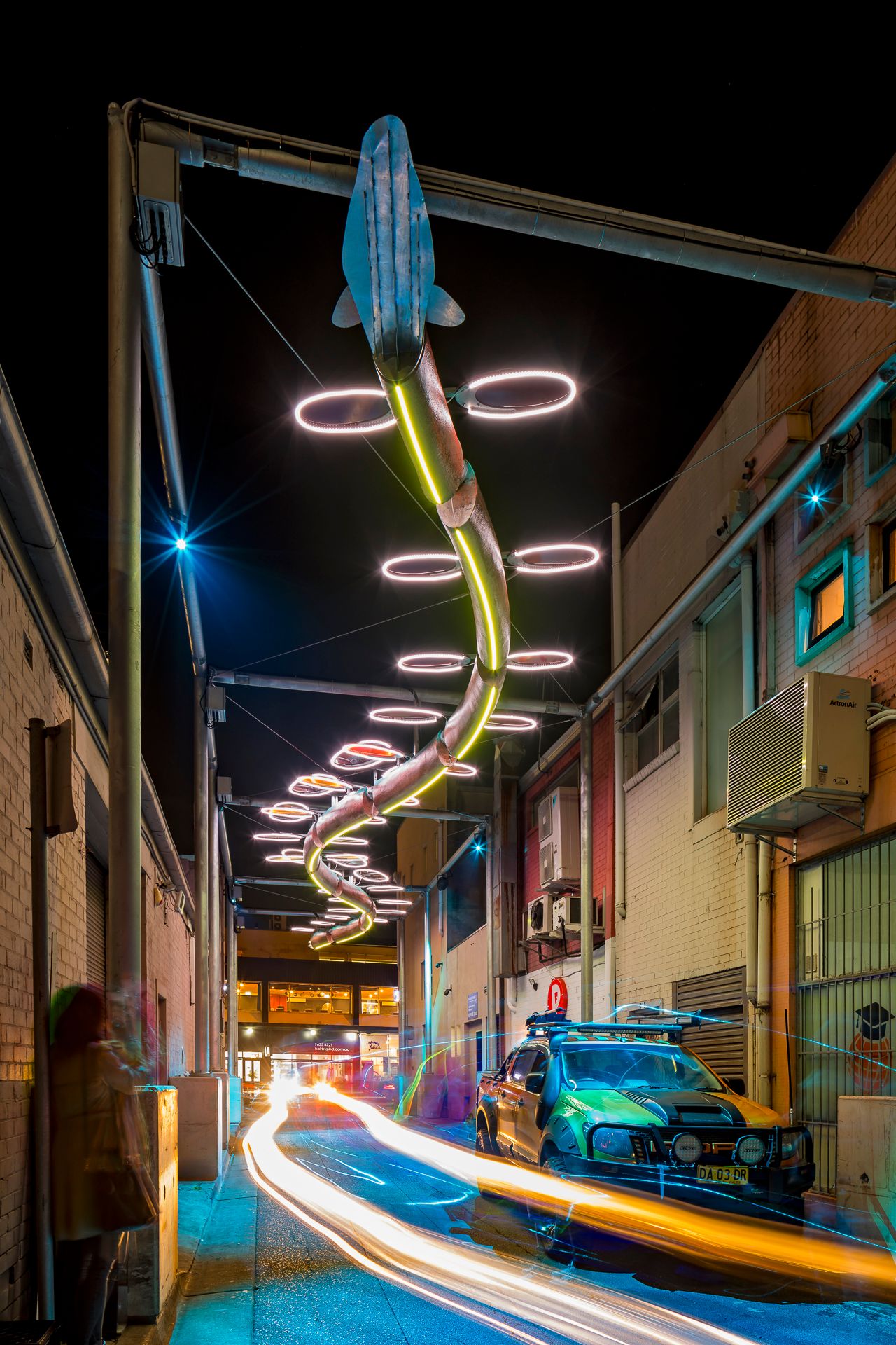 Glowing white and yellow neon fish installation above a night alley, with a parked utility vehicle and long-exposure light streaks.