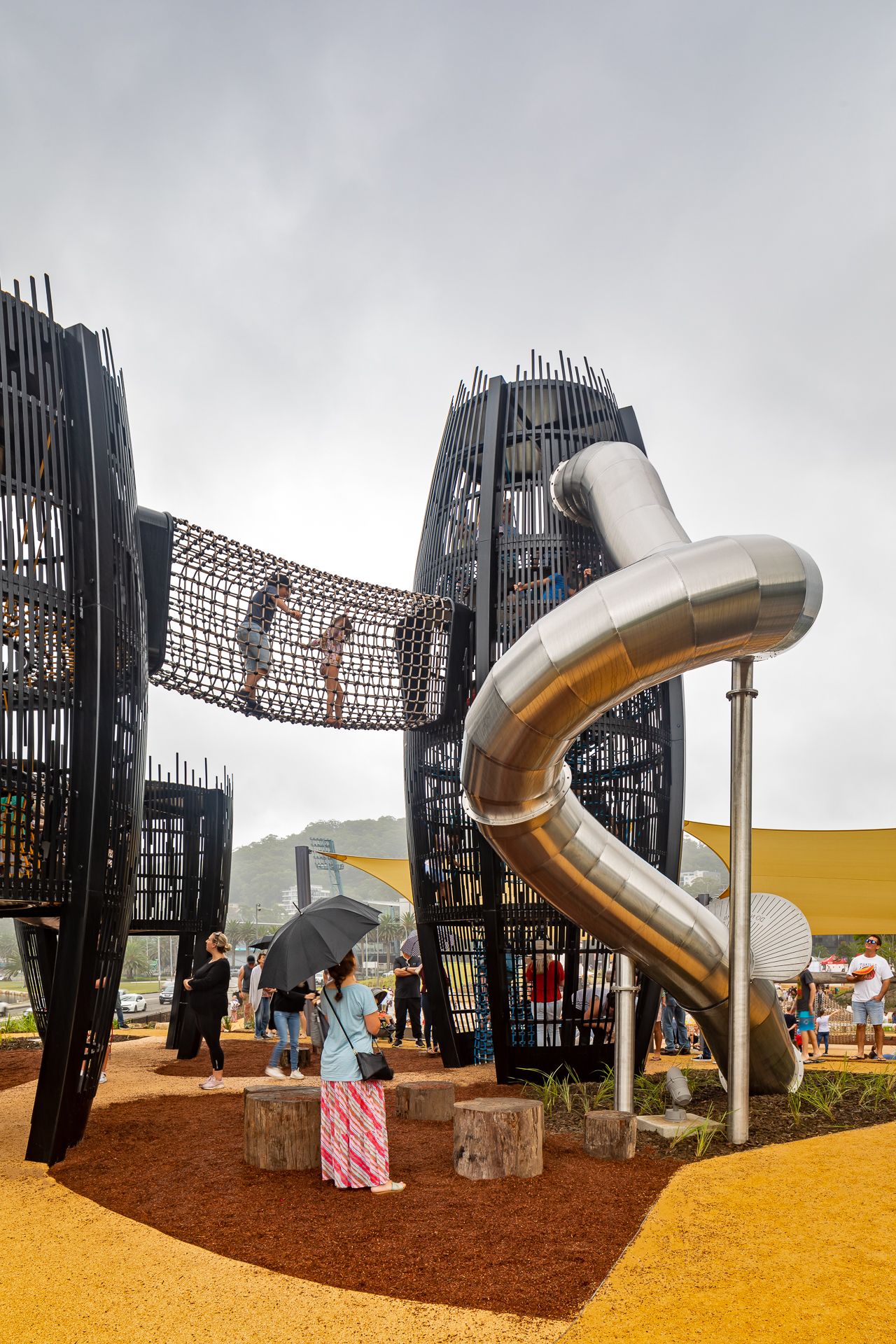 A parent with an umbrella watches children cross a rope net bridge between black climbing towers with a giant steel tube slide.