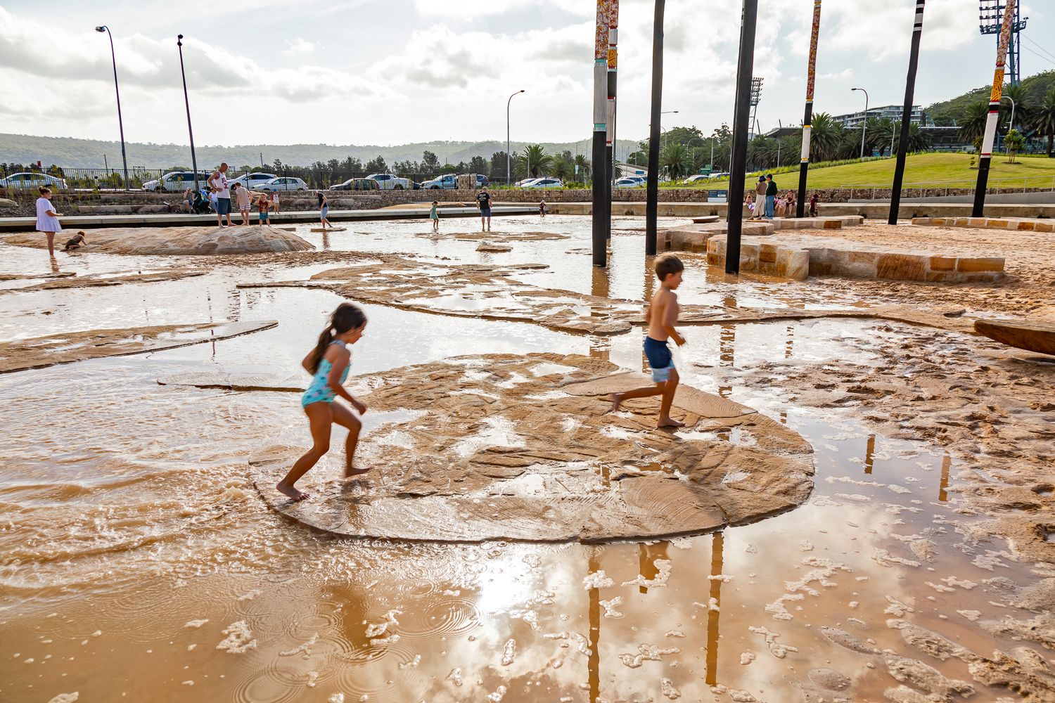 Two children run and splash across a shallow, muddy water play area in a sunny public park with hills in the background.