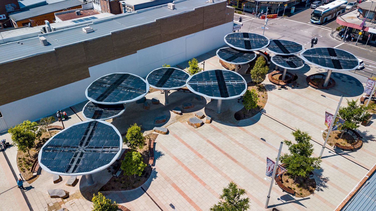 Aerial angle of round solar canopies over seating areas in a paved public square lined with planters and flags.