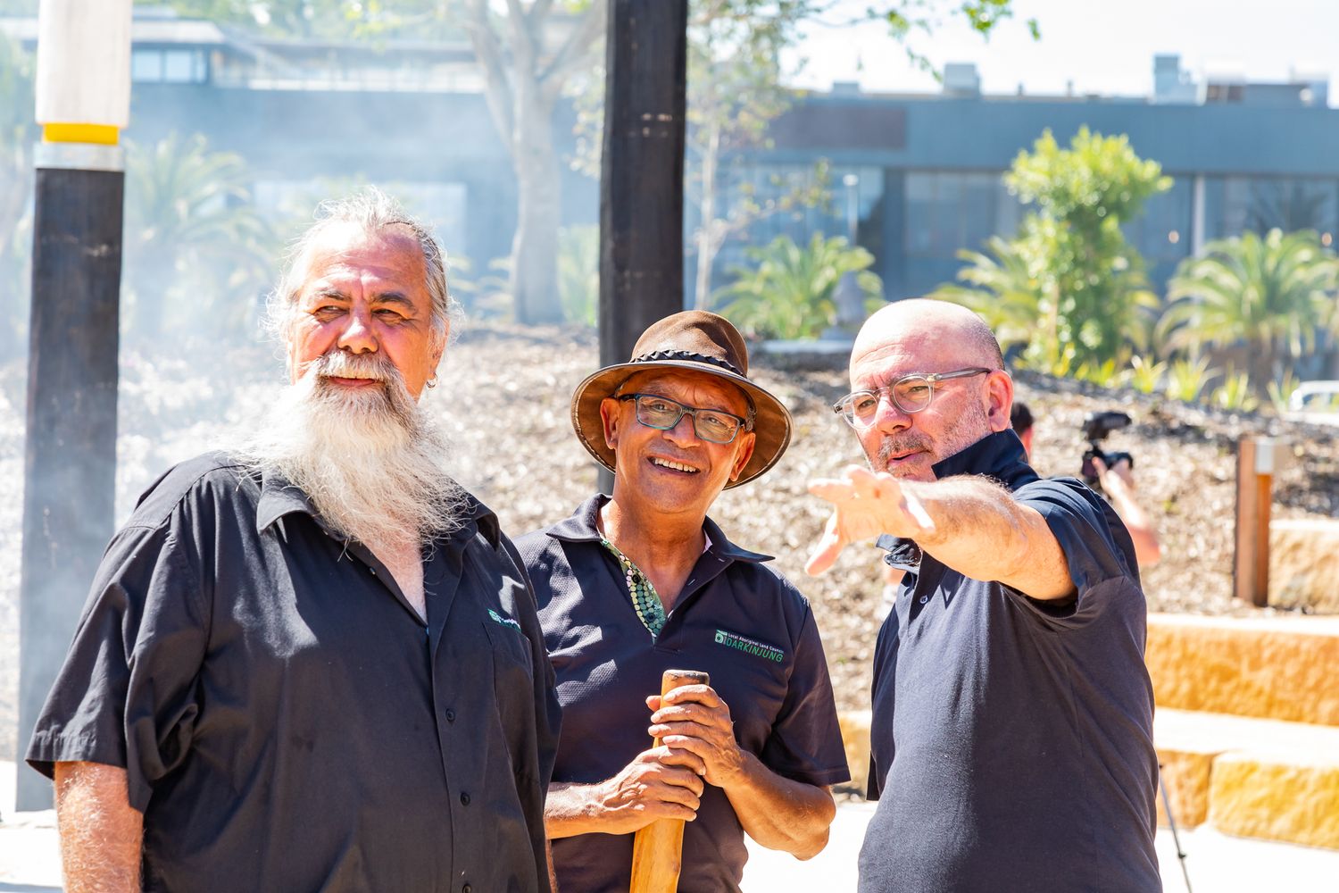 Three men chat outdoors at a sunny park, one with a long white beard and one in a brown hat, with a painted pole beside them.