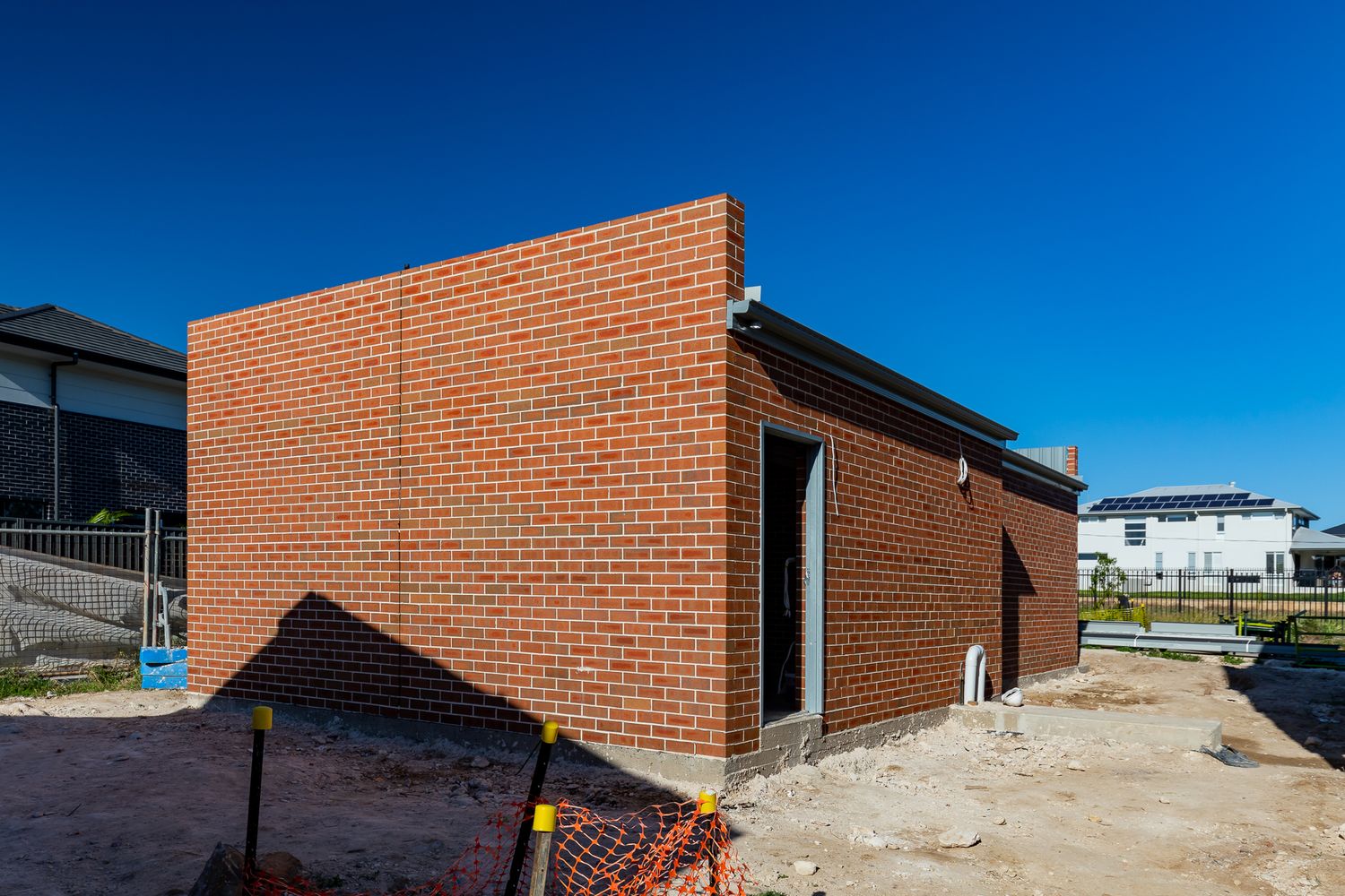 Single-storey red brick structure under construction on a sandy building site beneath a clear blue sky.