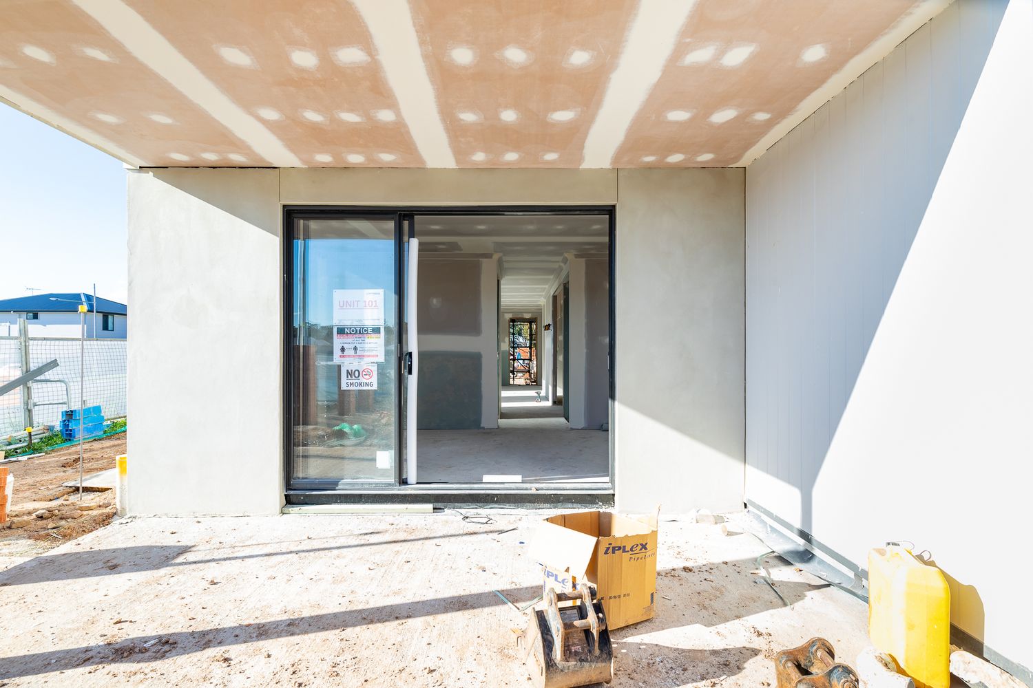 Newly built townhouse under construction with sliding glass doors, an exposed ceiling and building materials on the dirt ground outside.