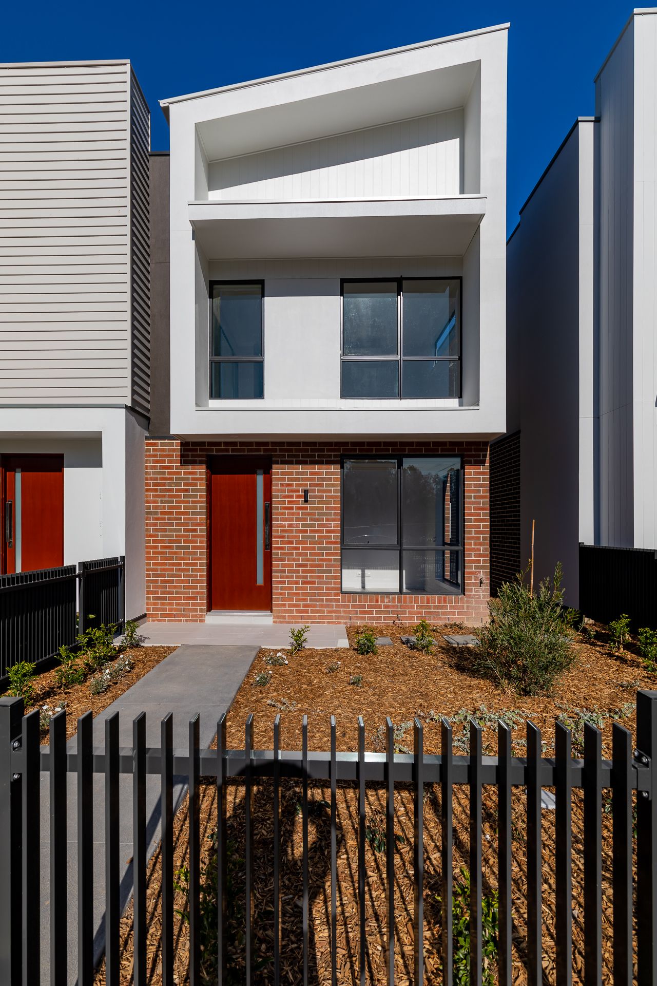 Modern two-storey townhouse with white upper facade, red brick base, red front door and a black picket fence with landscaped garden.