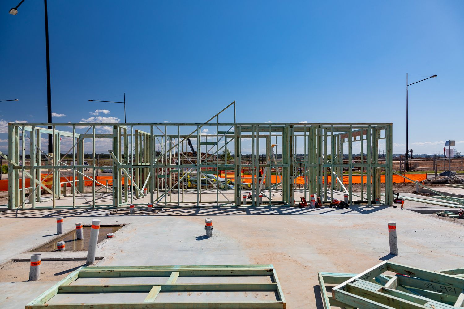 Timber wall frames standing on a concrete slab at a construction site, with light poles and a bright blue sky in the background.