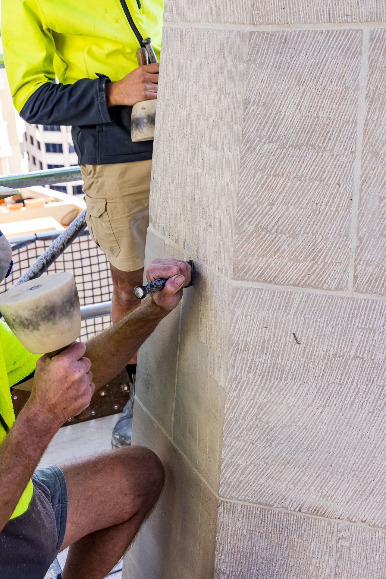 Stonemason in high-visibility clothing uses a mallet and chisel to carve textured sandstone masonry.