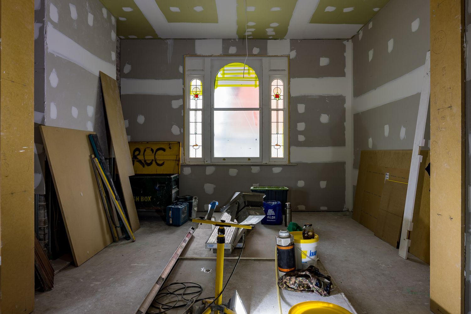 Room under renovation with unfinished drywall, arched stained-glass window, building materials, and tools on a dusty floor.