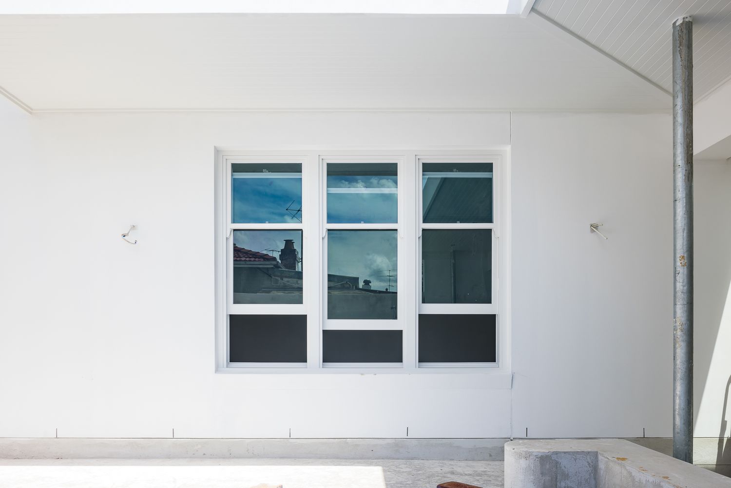 White rendered exterior wall with a modern triple window with white frames reflecting the sky, beside a metal downpipe.