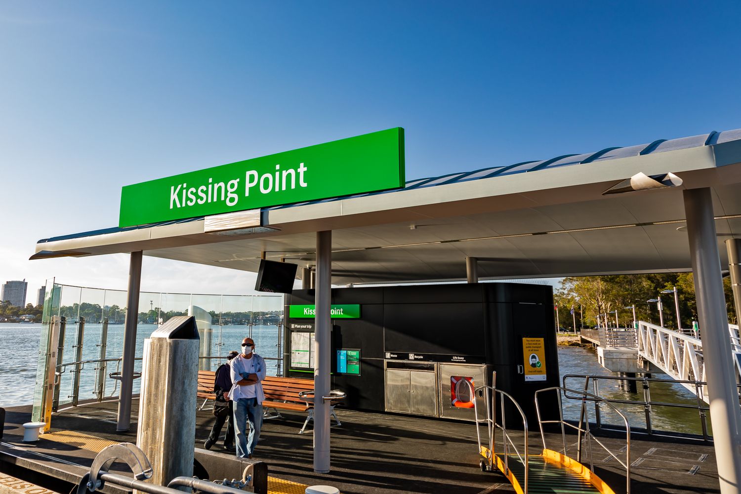 Commuters wait under the canopy at Kissing Point ferry wharf beneath a green station sign, with the water behind them.